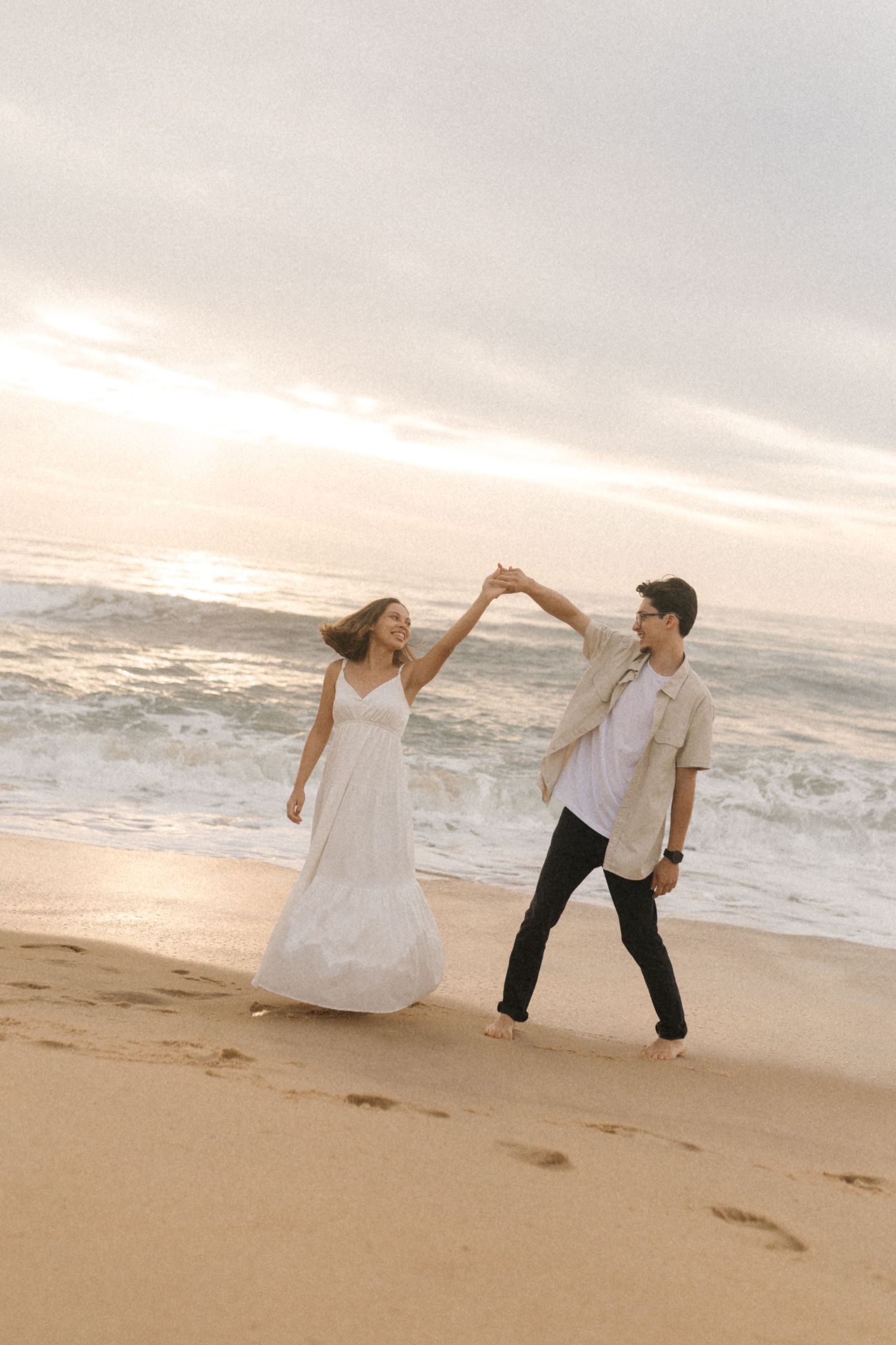 A couple dances together on a sandy beach at sunset, with the bride wearing a flowing white dress.