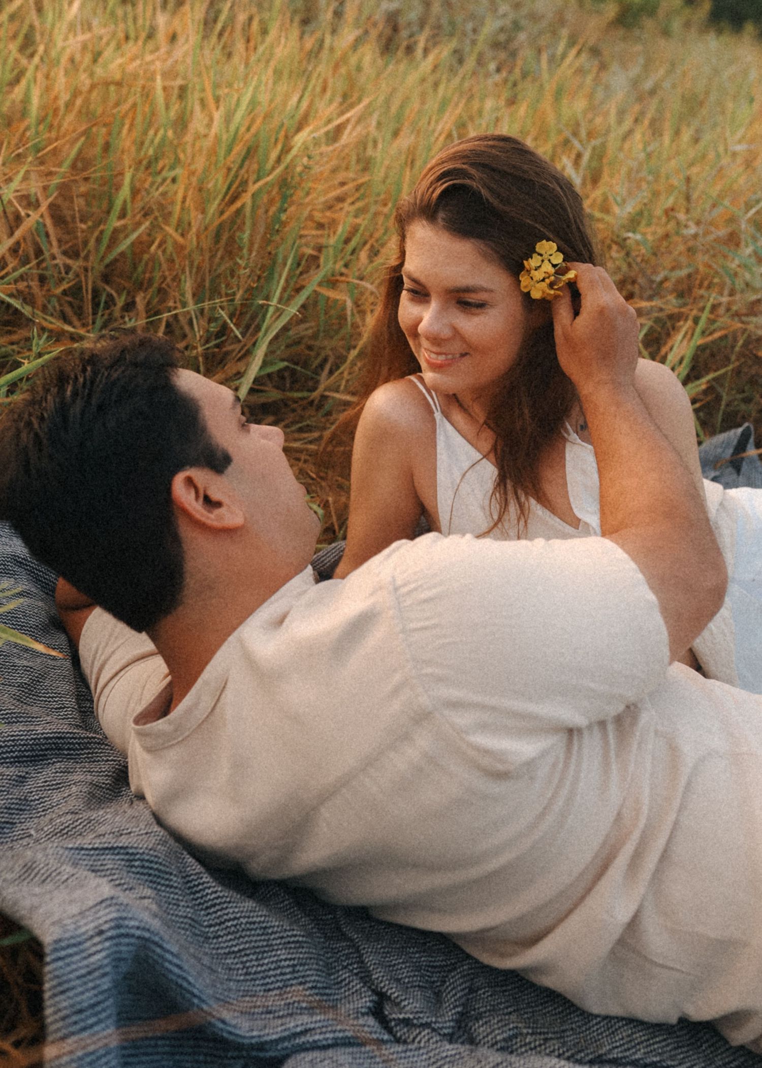Couple lying on blanket in tall grass sharing an intimate moment in white clothing.