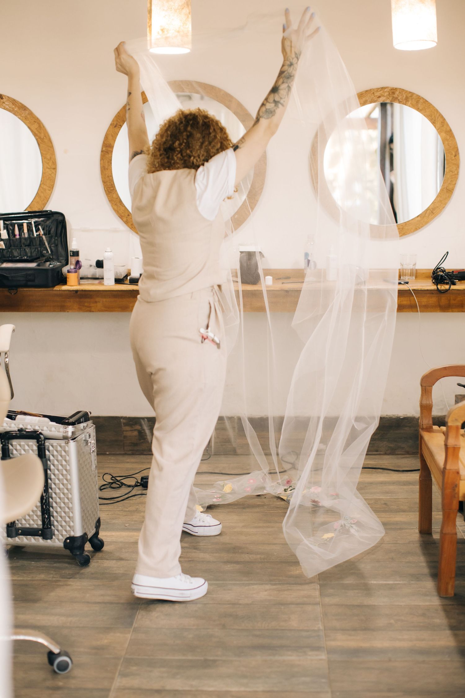 Stylist celebrating with raised arms while working with wedding veil in salon with circular mirrors.
