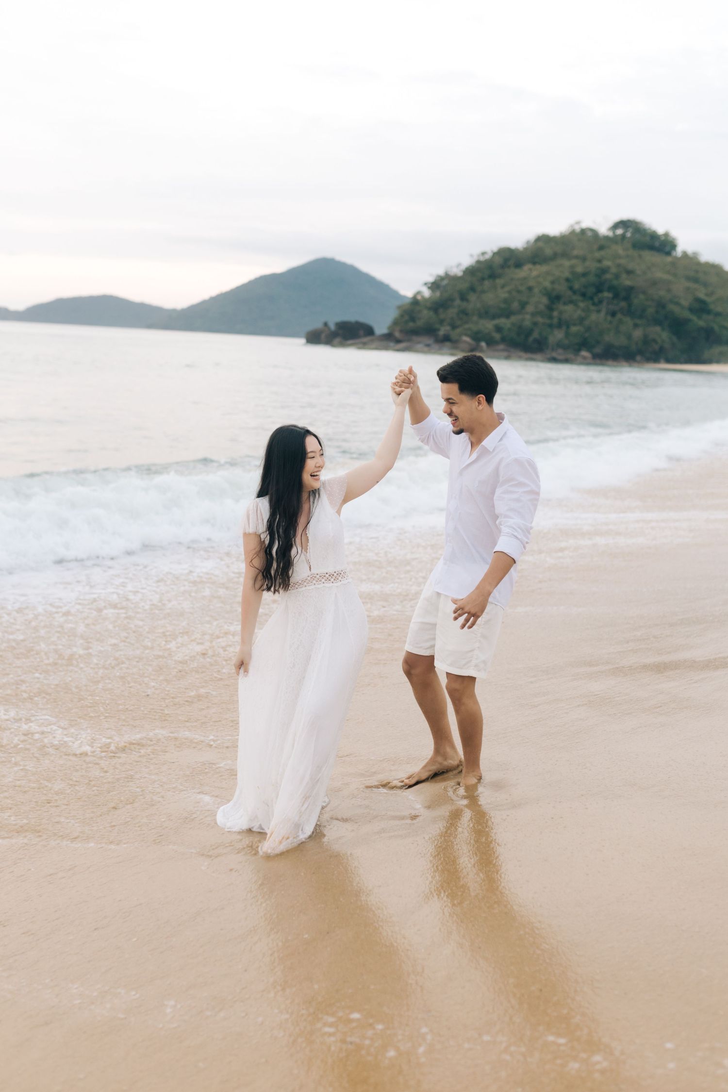 A couple dances together on a scenic beach with mountains in the background.