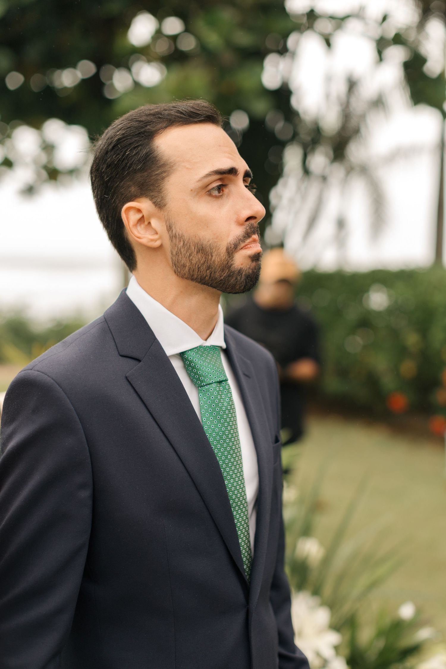 A man in a dark suit with green tie looks thoughtfully into the distance at an outdoor event.