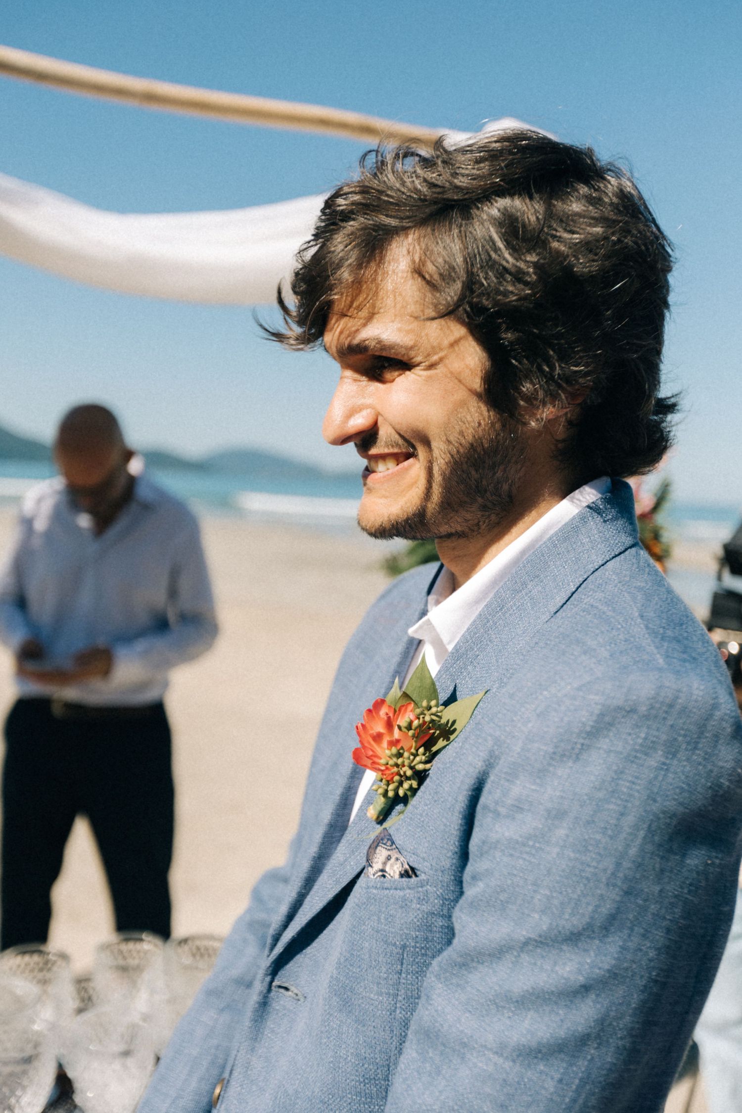 A groom smiles during an emotional beach wedding ceremony while wearing a blue suit.