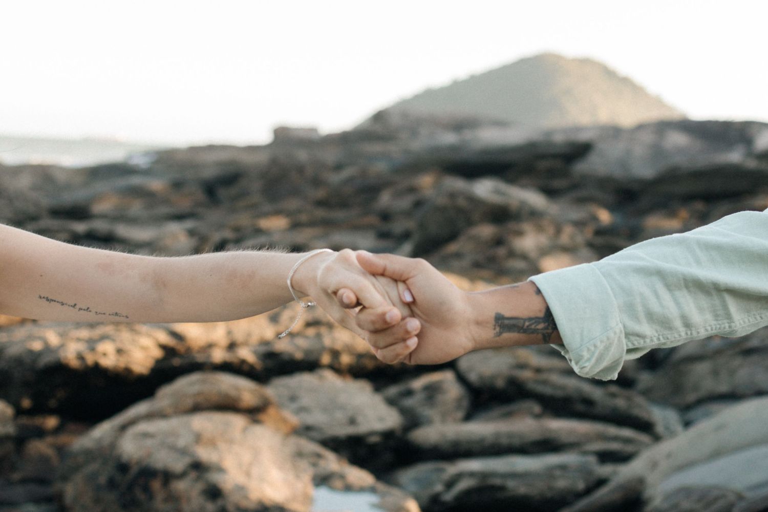 Close-up shot of two hands clasped together against a rocky volcanic landscape at golden hour.