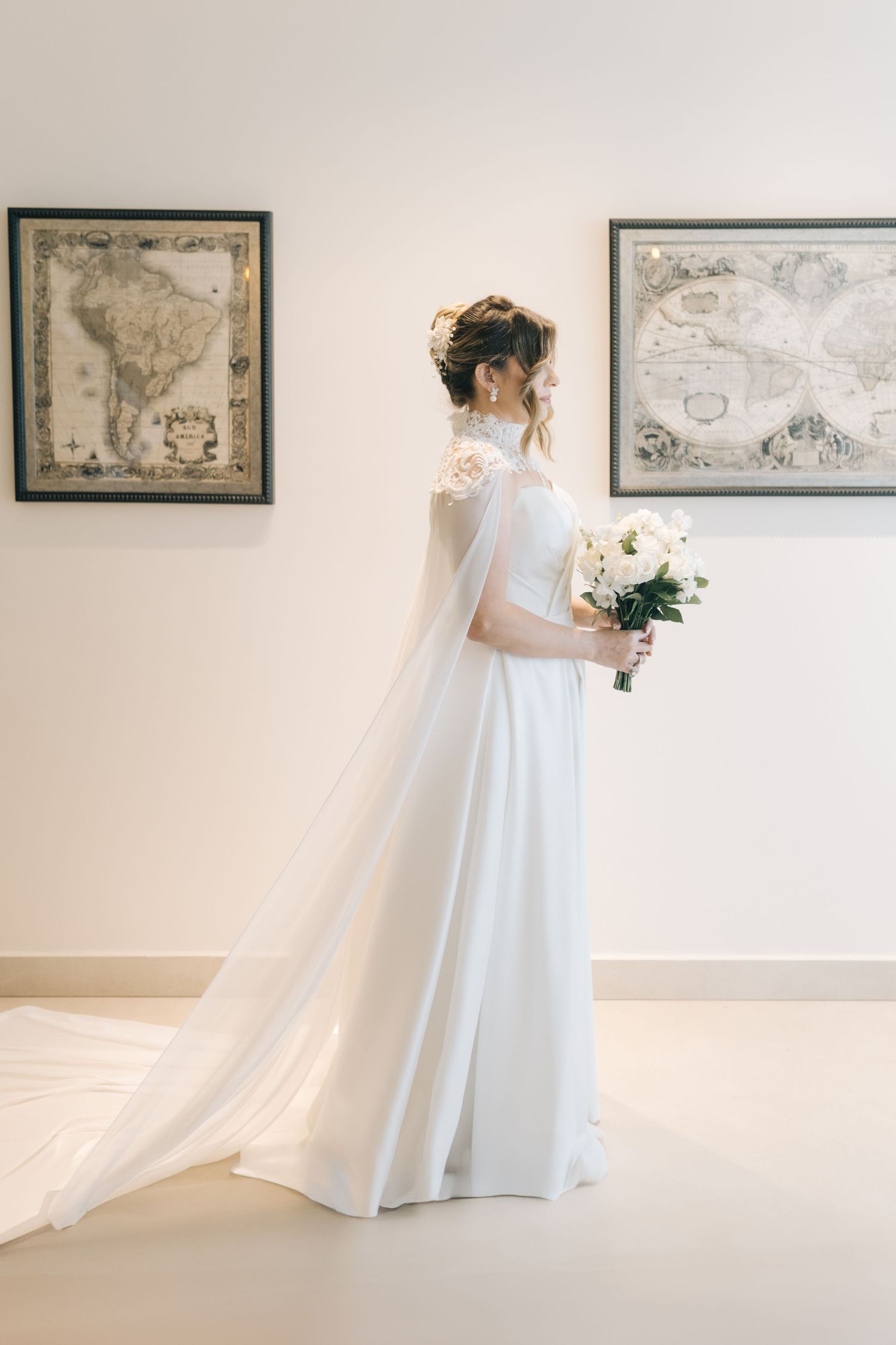 An elegant bride in a flowing white dress stands before vintage map artwork holding a white bouquet.