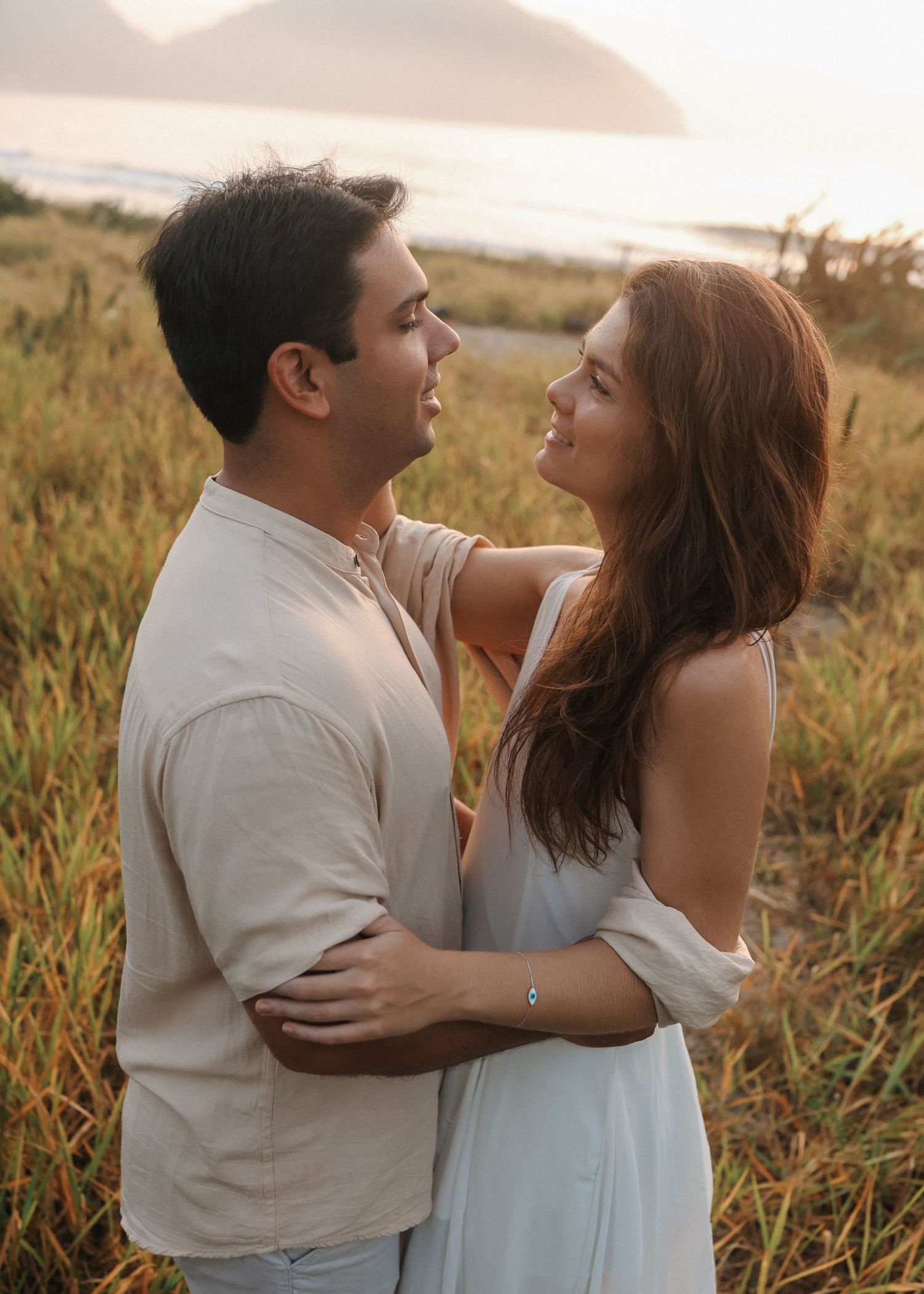 A couple in white clothing laughs together in a scenic field during golden hour.