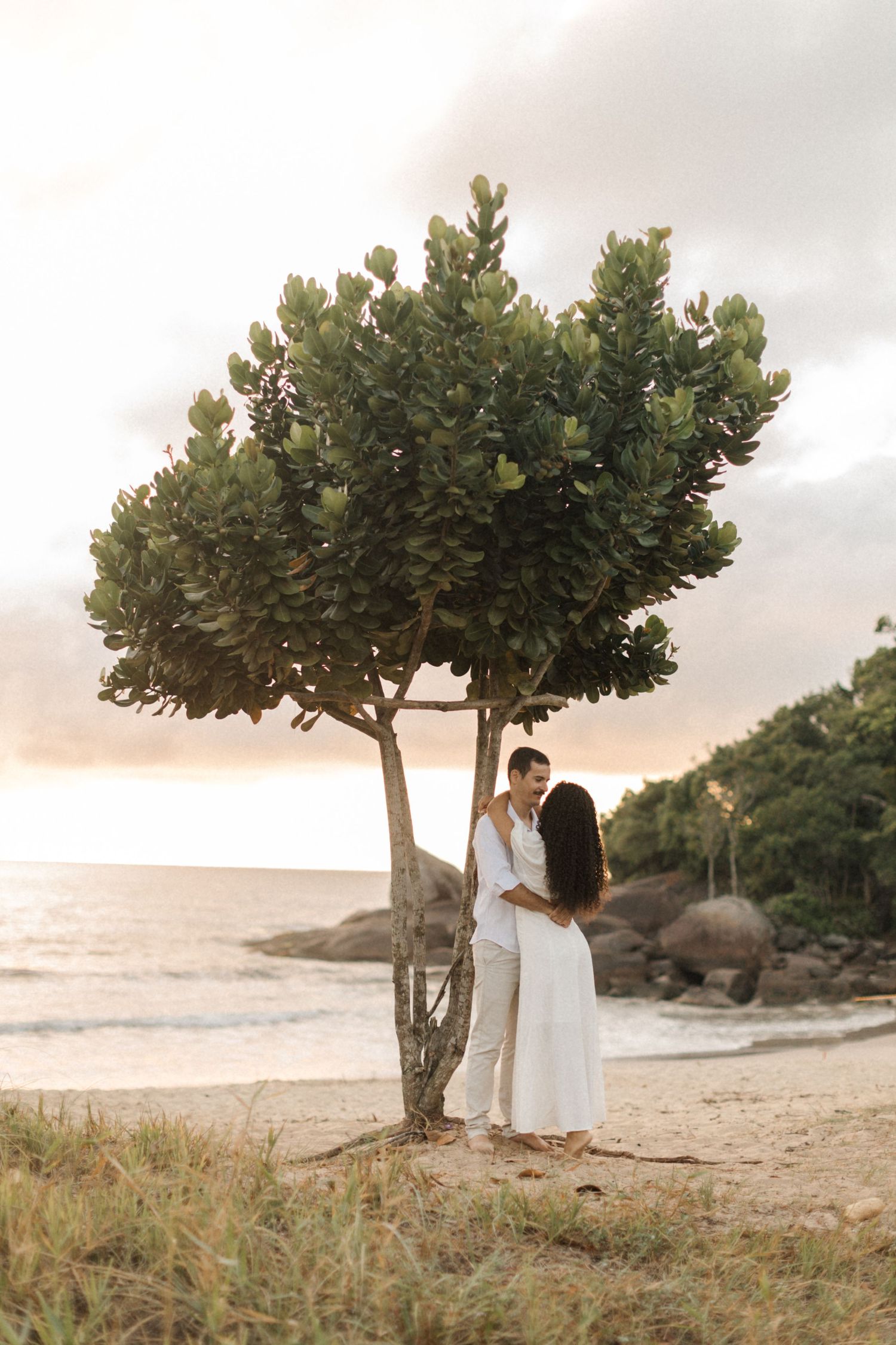 A couple in white shares an intimate moment beneath a tropical tree on a sandy beach at sunset.