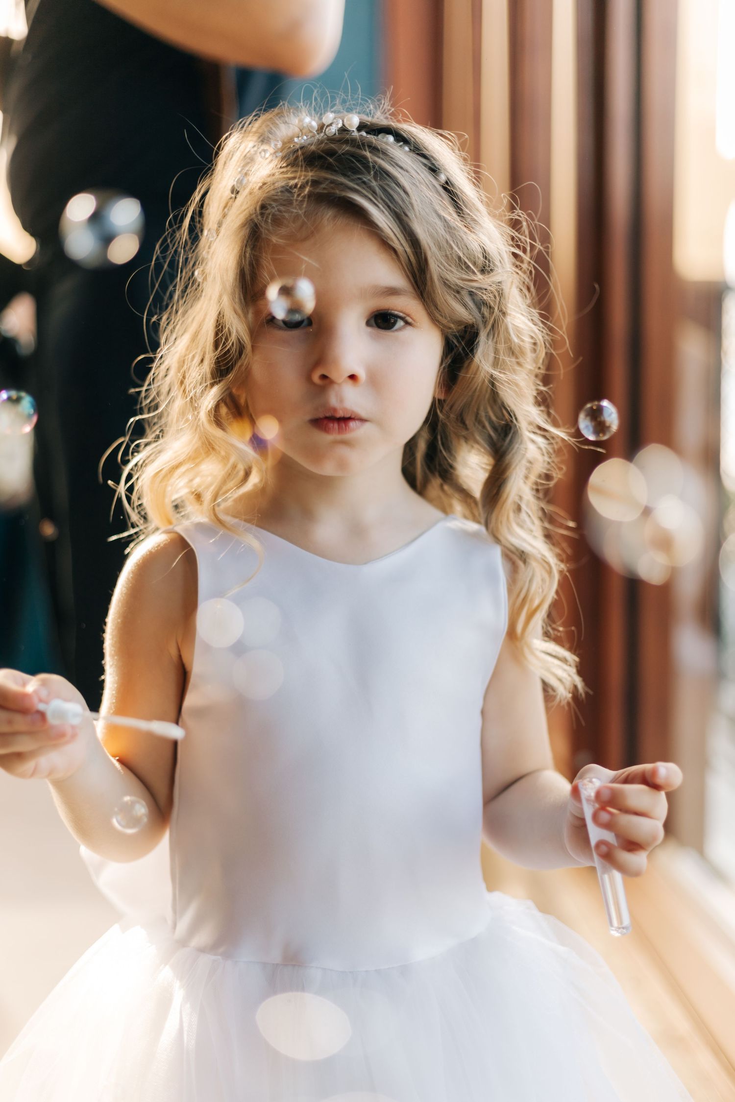 Young flower girl in white dress playing with bubbles at wedding celebration.