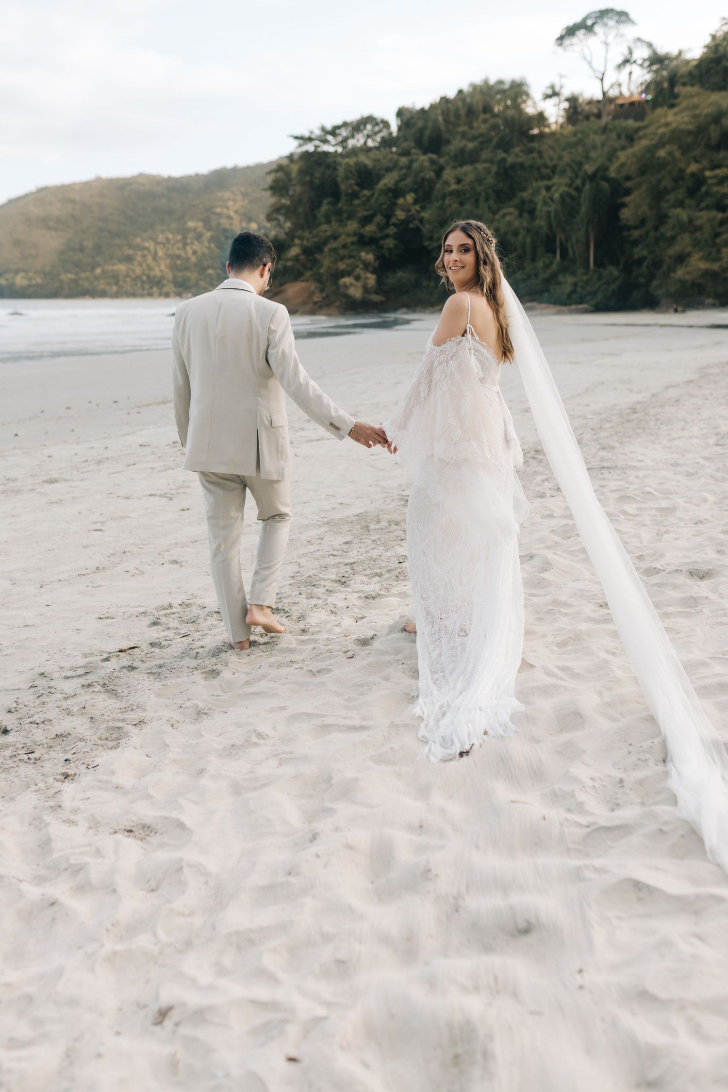 Couple walking hand in hand along pristine beach with flowing white veil trailing behind.