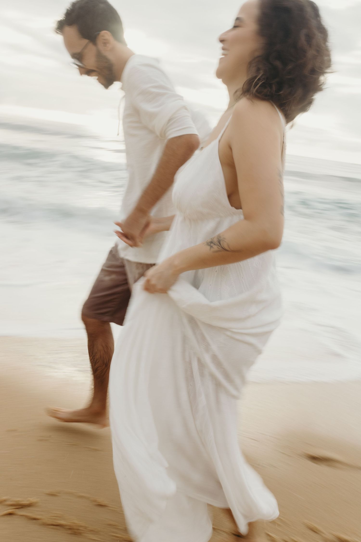 A couple in white clothing dances on a sandy beach with ocean waves behind them.