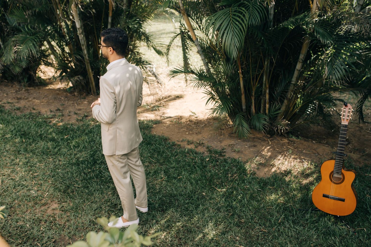 A person in a light colored suit stands in a tropical garden setting with an acoustic guitar visible.