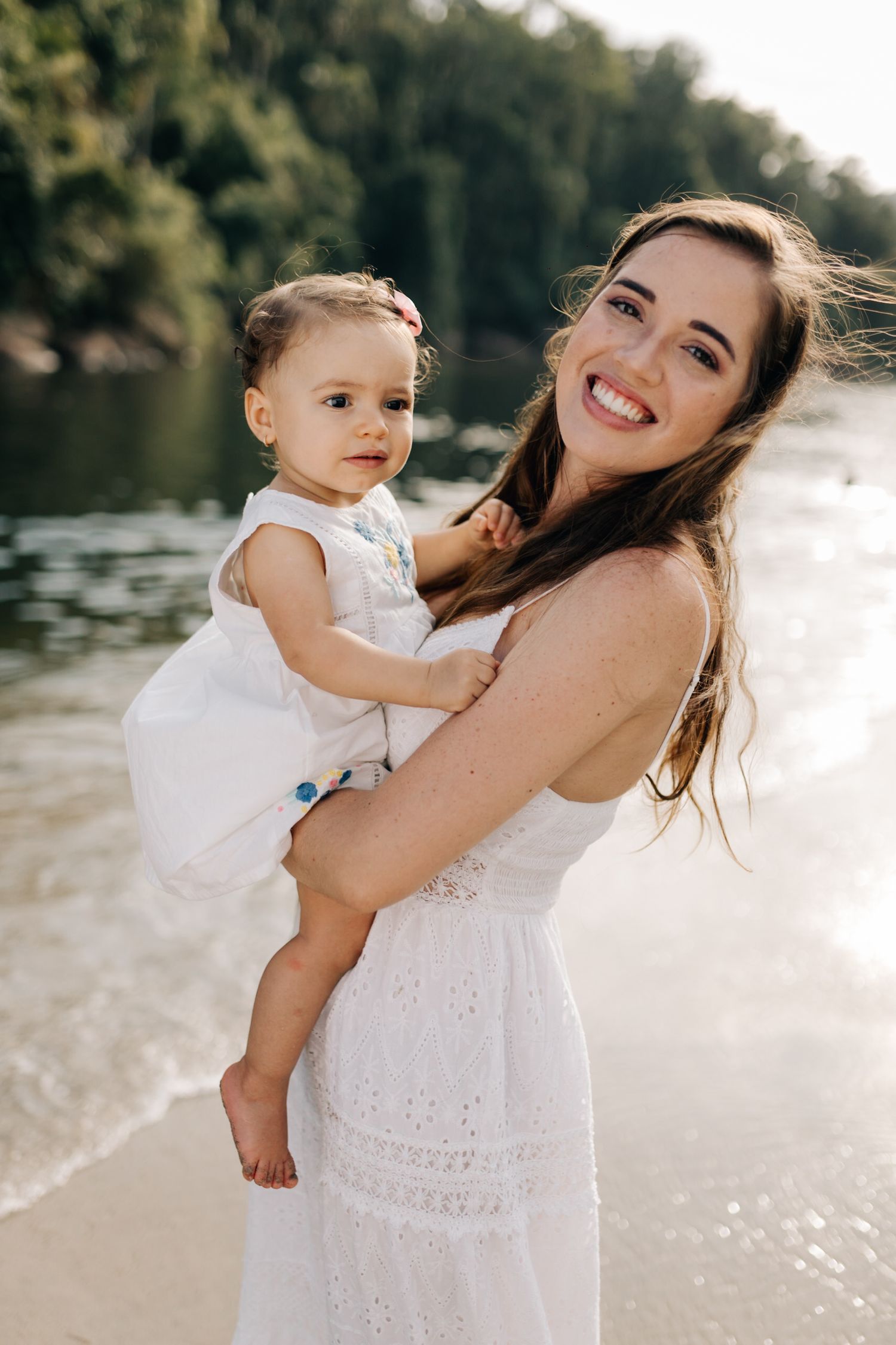 Joyful outdoor portrait of parent and child in white dresses by a lake.
