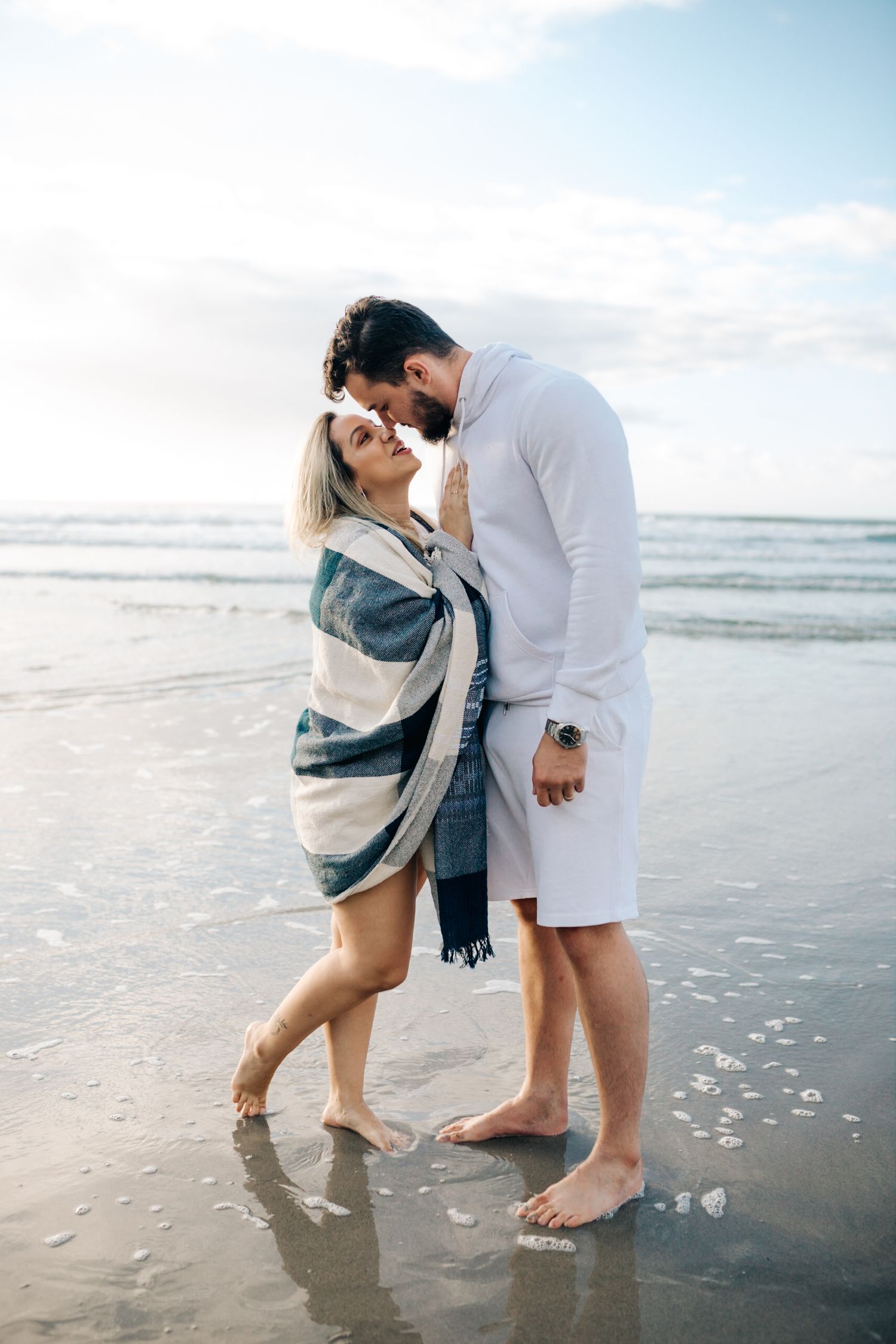 Intimate beachside moment between couple wrapped in patterned blanket at waters edge.