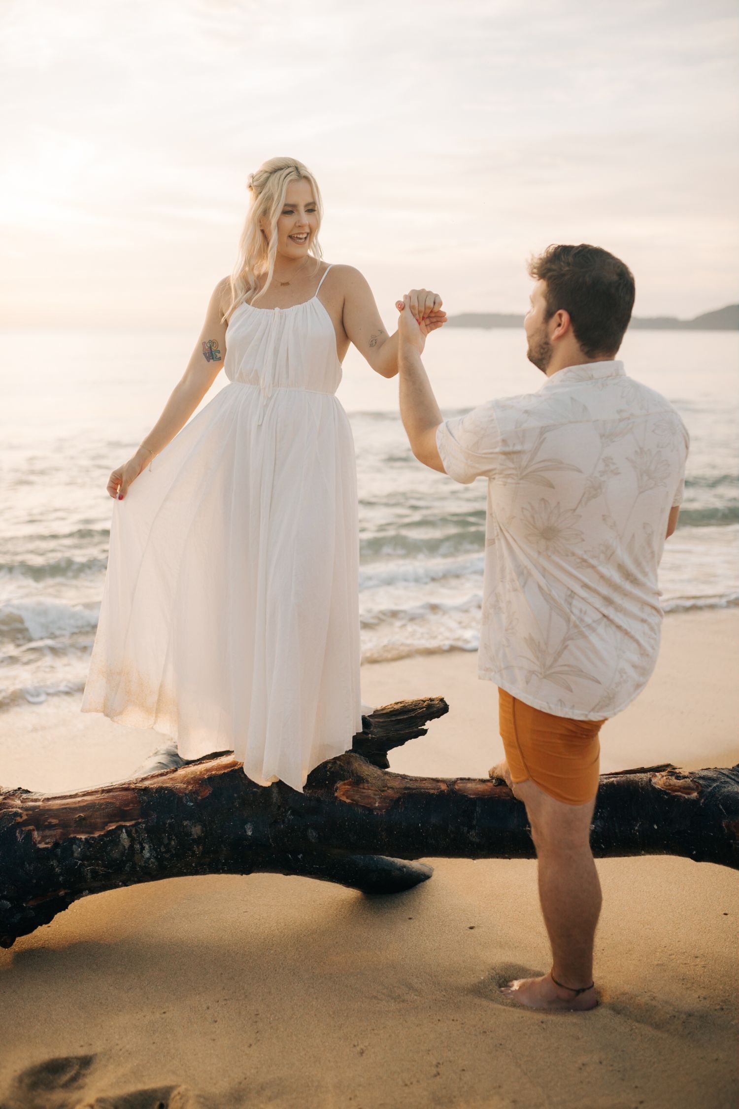 A couple holds hands while balancing on a fallen tree trunk at the beach.