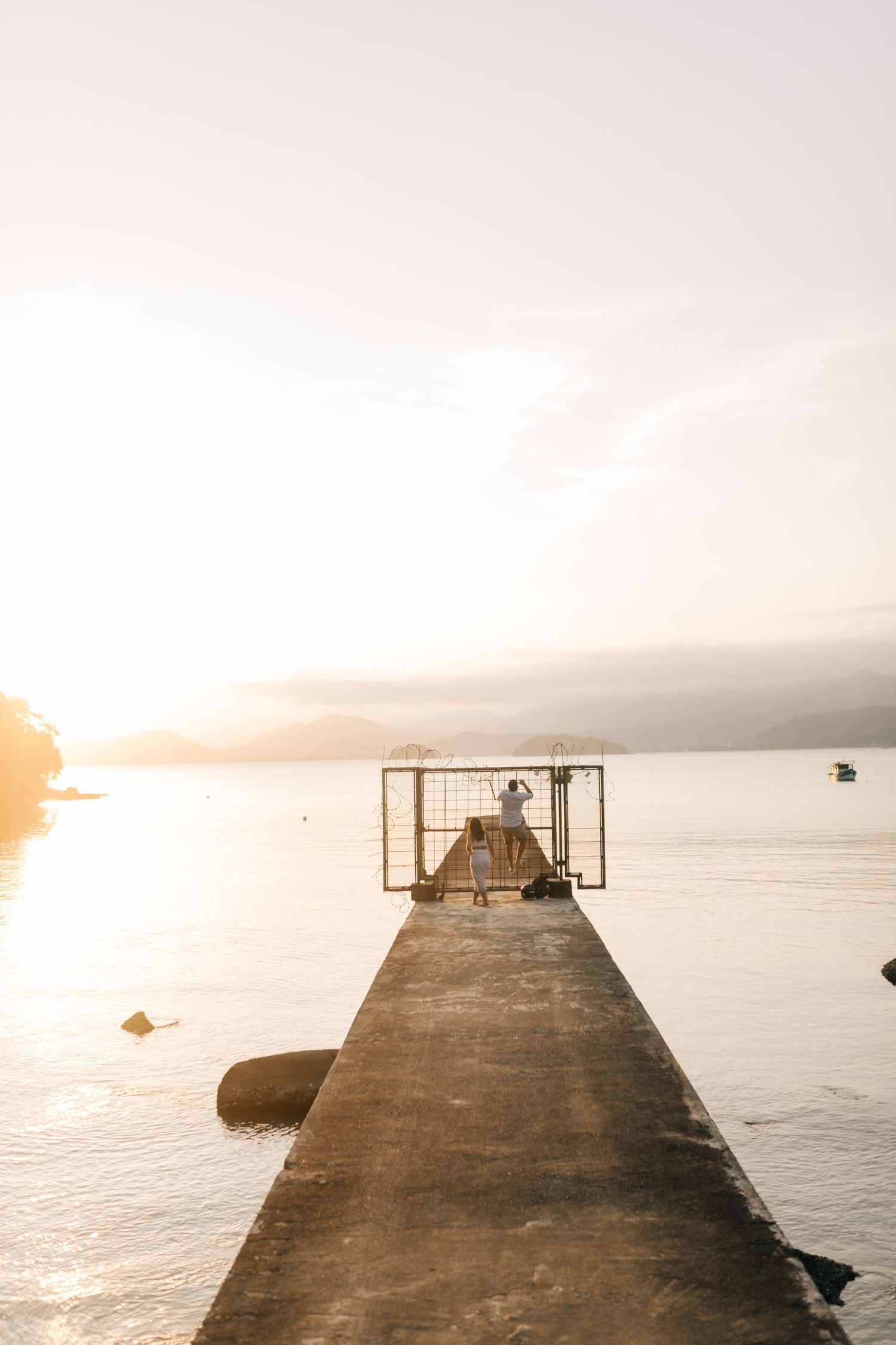 Long concrete pier extending into calm water at sunset with metal structure at end.