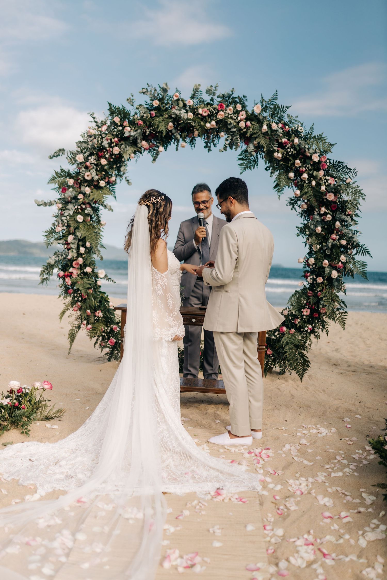 A gorgeous floral arch frames the wedding ceremony on a scenic beach with scattered rose petals.