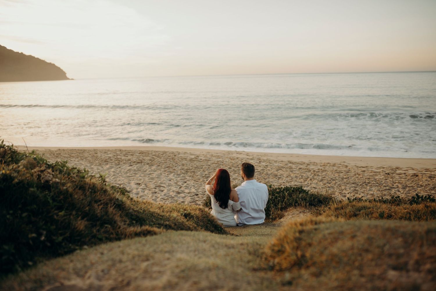 Two people sit together on a coastal cliff overlooking the ocean at sunset.