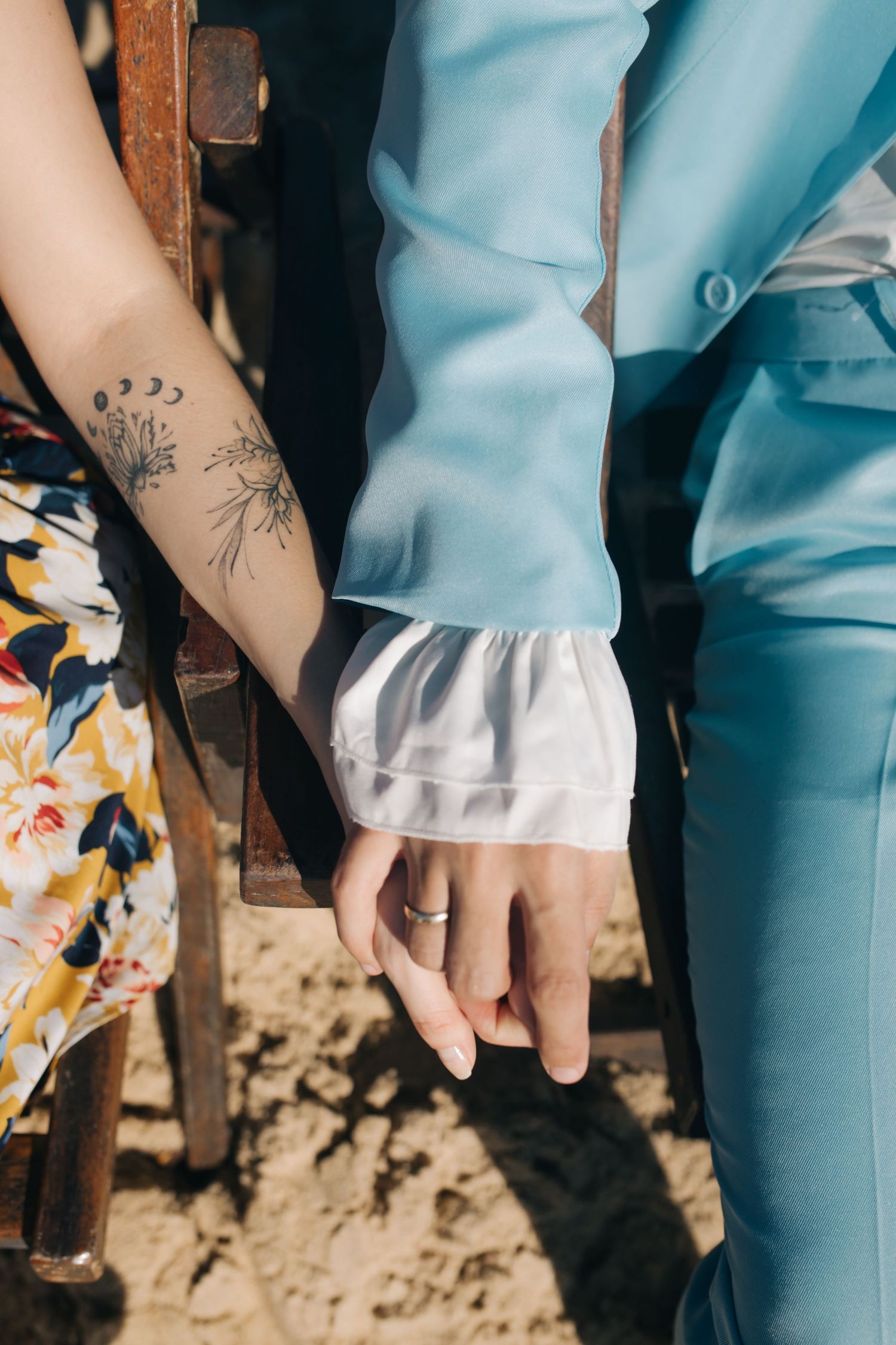 Close-up of hands and floral dress details against light blue suit on beach.