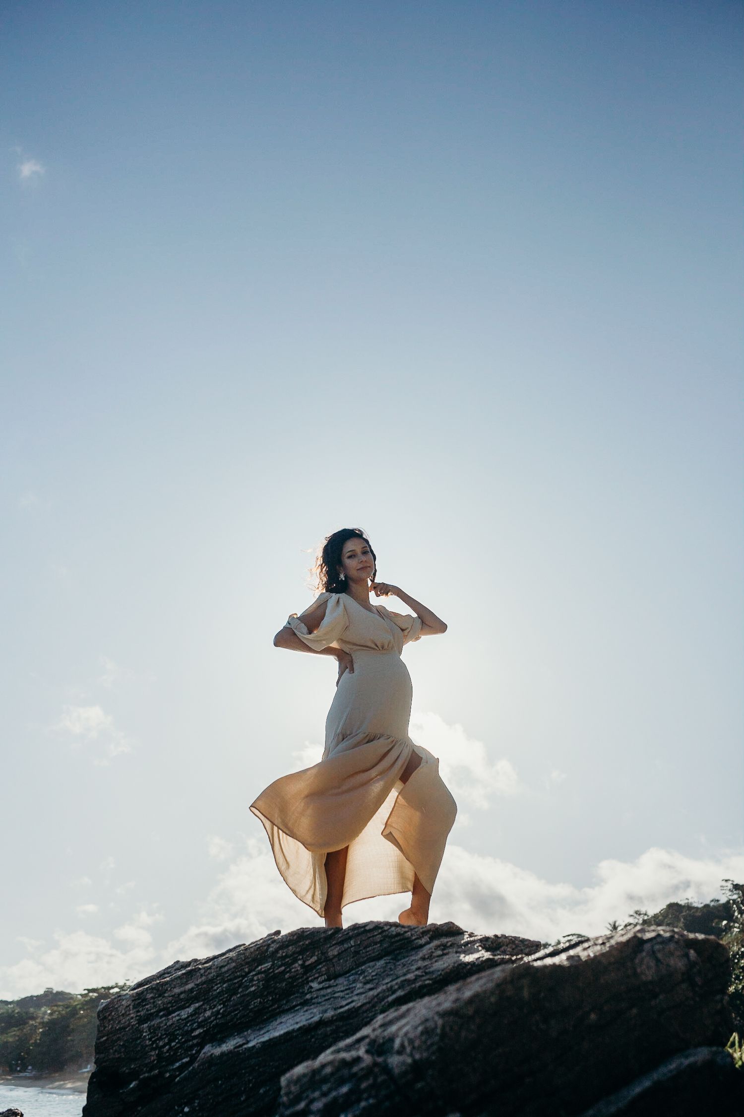 Figure in light dress poses gracefully on rocky cliff with ocean backdrop.