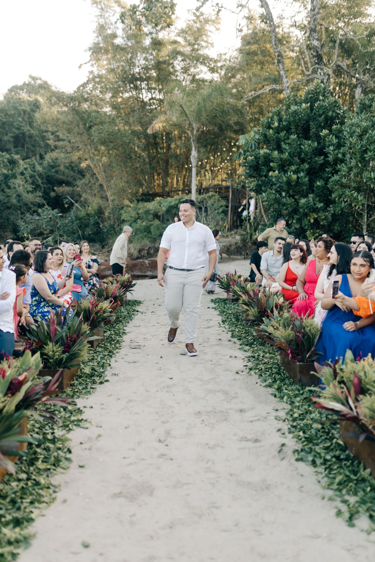 Outdoor wedding ceremony setup with guests seated along a sandy aisle bordered by tropical plants.