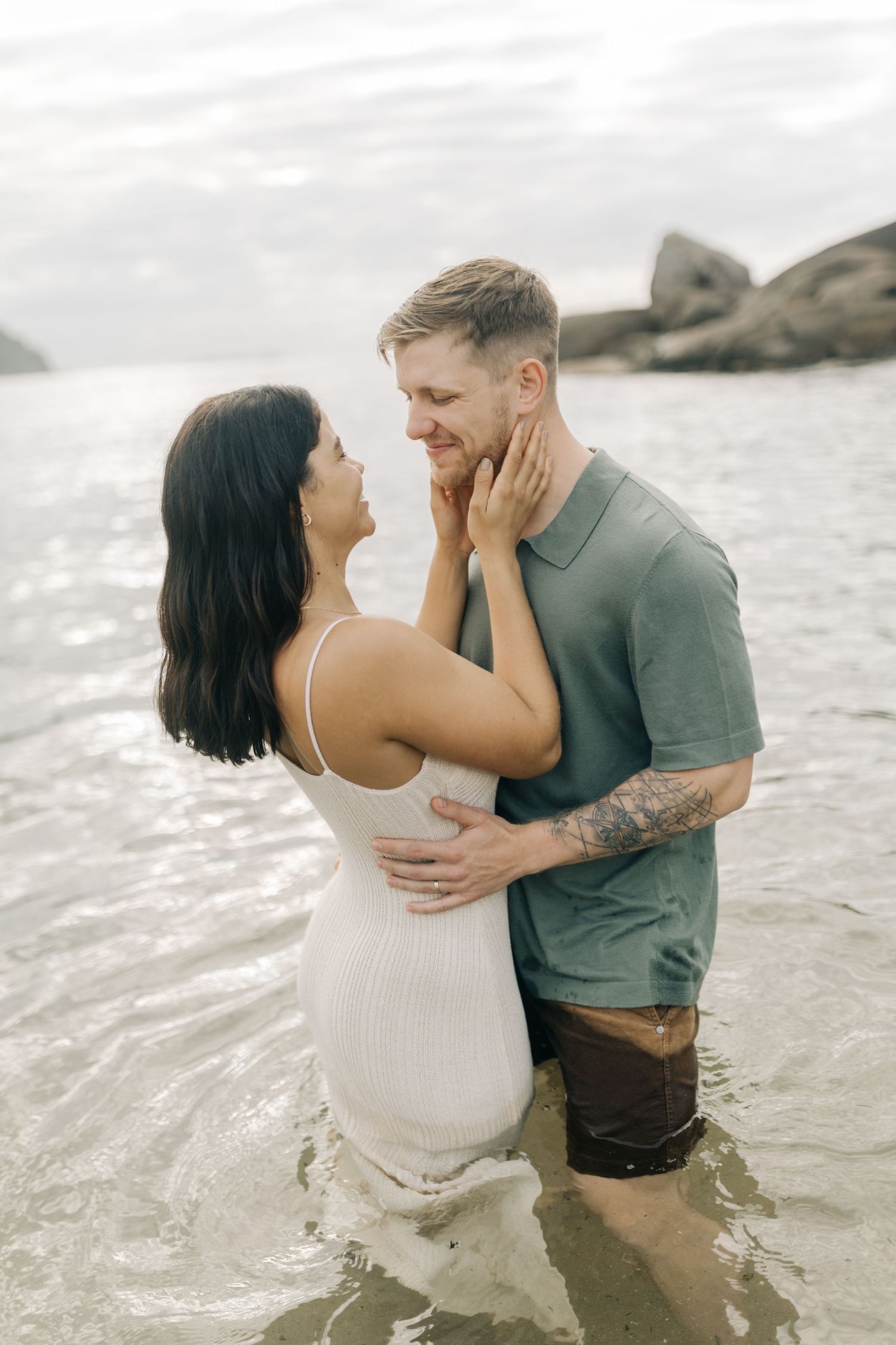 A romantic couple embraces while standing in shallow ocean water against a misty coastal backdrop.