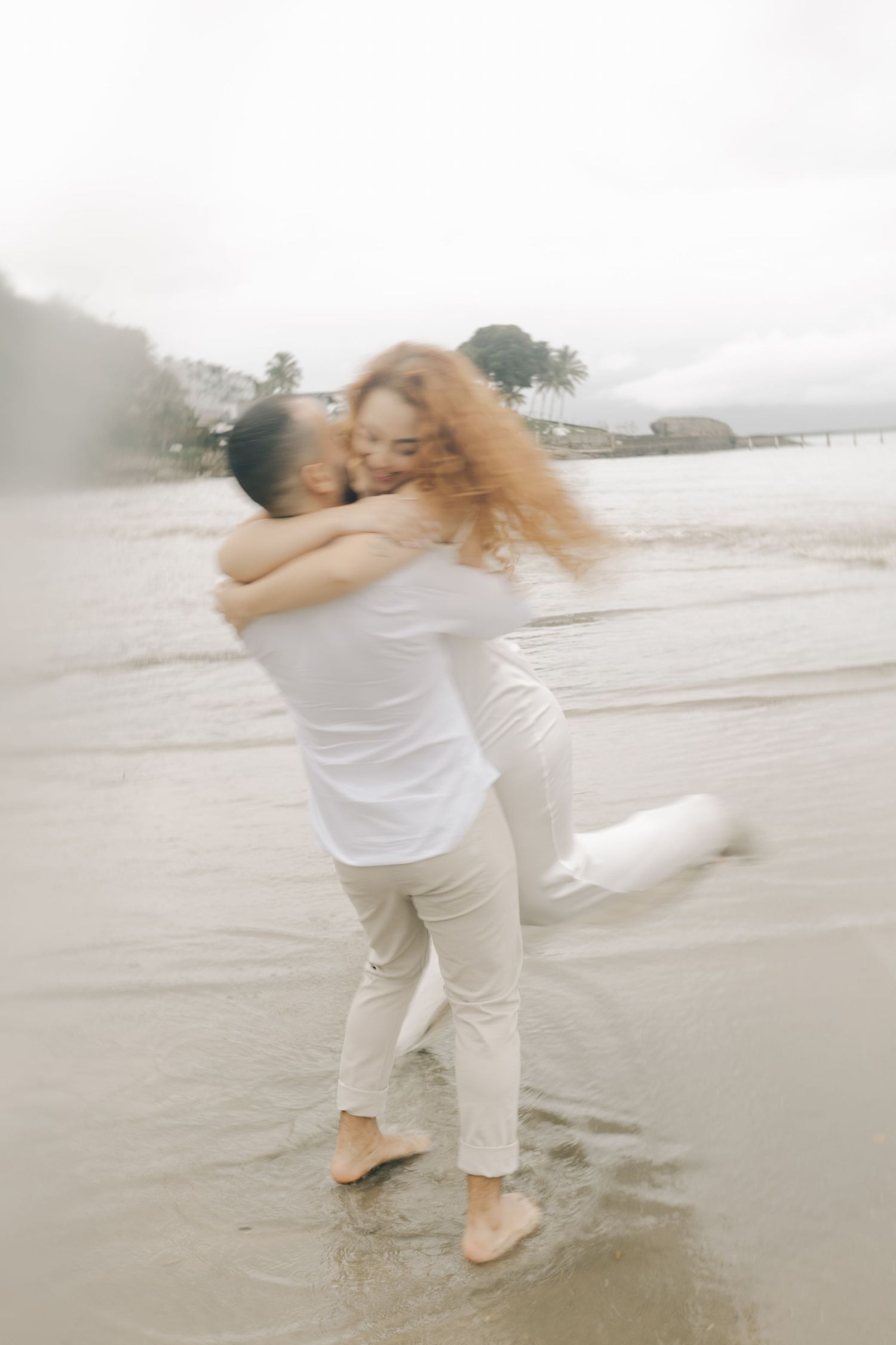 A couple in white attire shares a playful spinning dance moment on the beach.