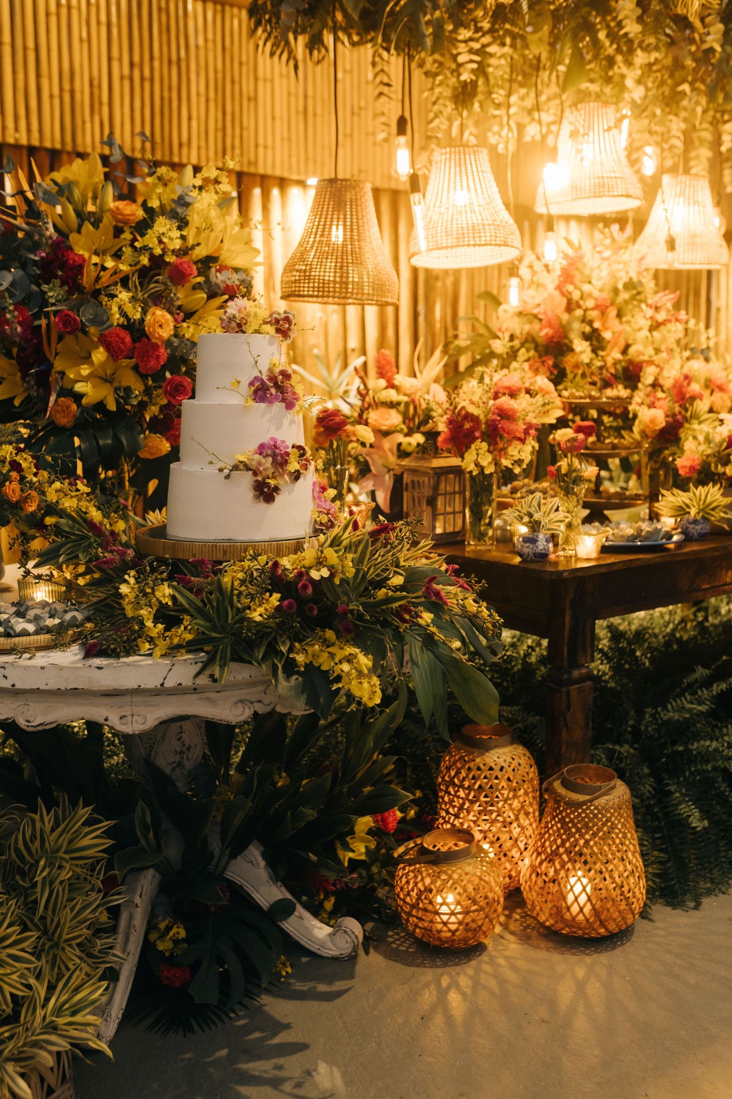 Decorative wedding table with gold lanterns, white wedding cake, and colorful floral arrangements in warm lighting.