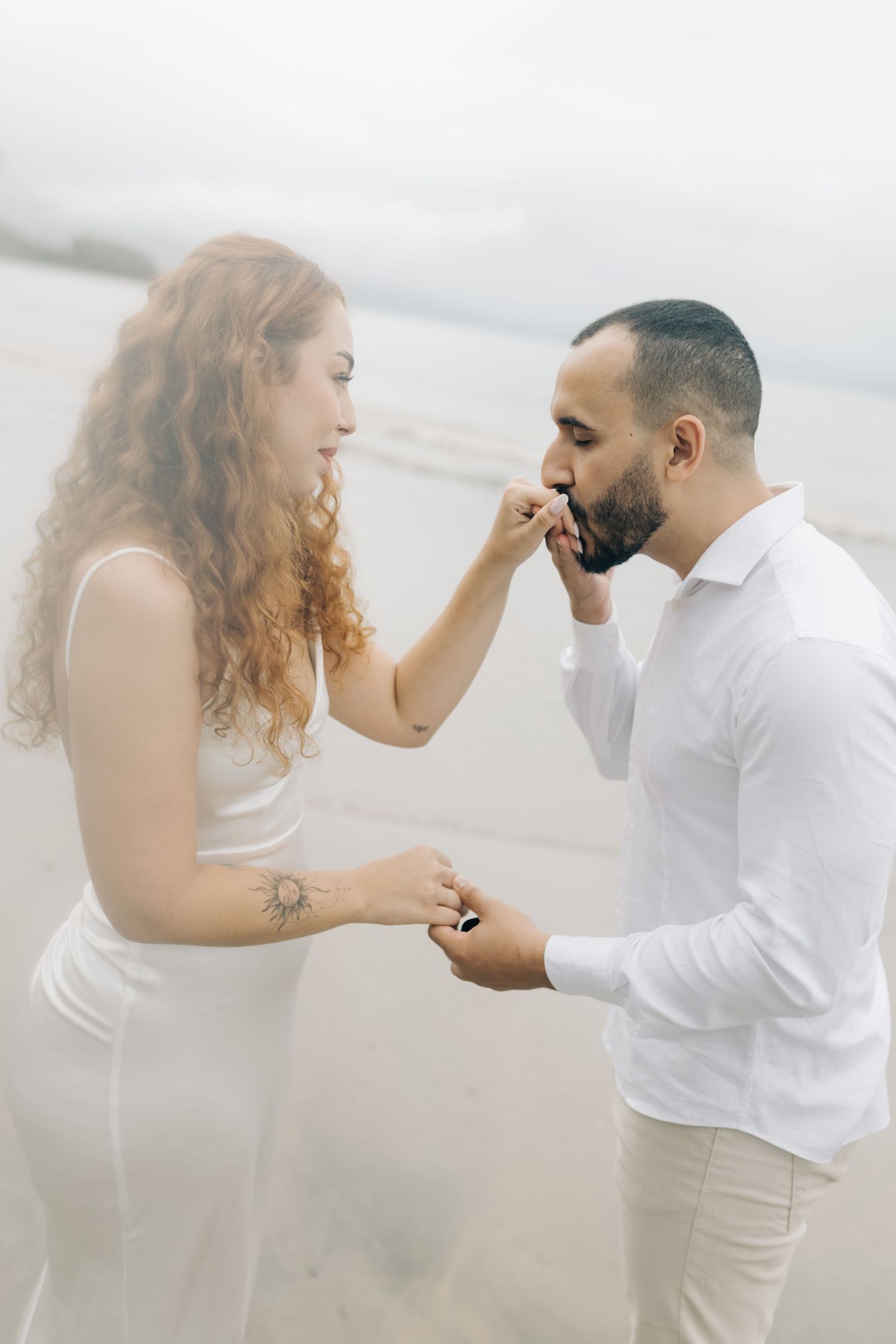 Romantic couple in white attire shares tender engagement moment on foggy beach.