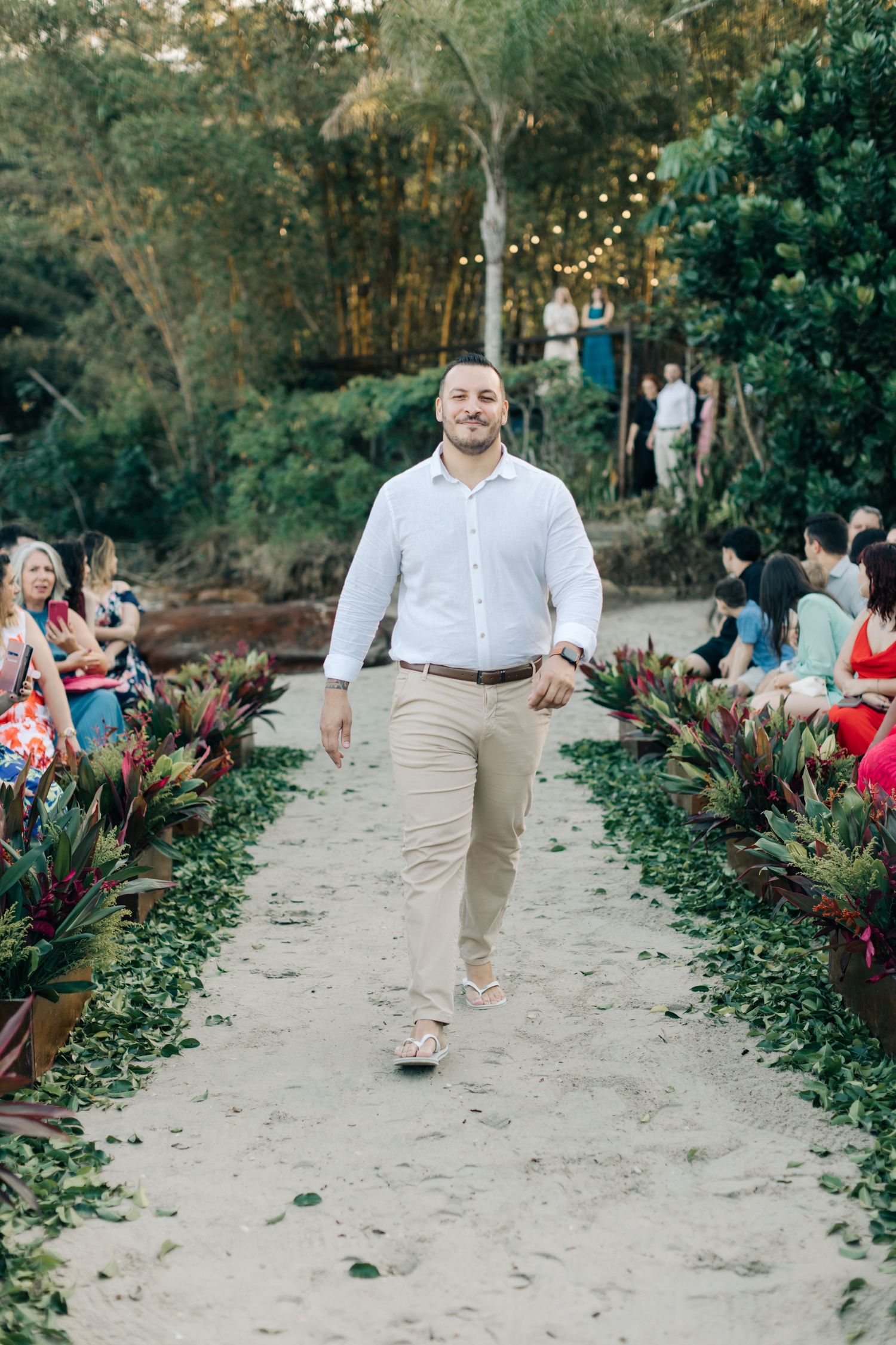 Wedding party member in white shirt and khakis walks down outdoor garden aisle.