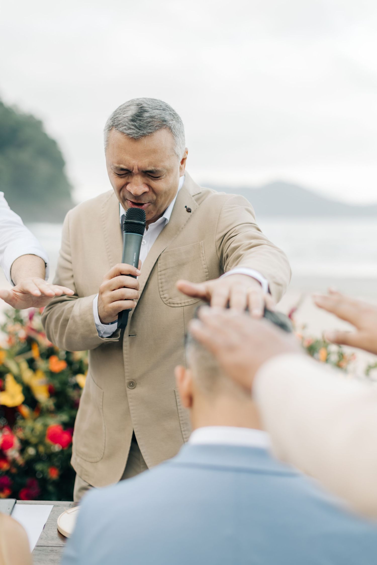 Person in beige suit speaking into microphone during beach wedding ceremony.
