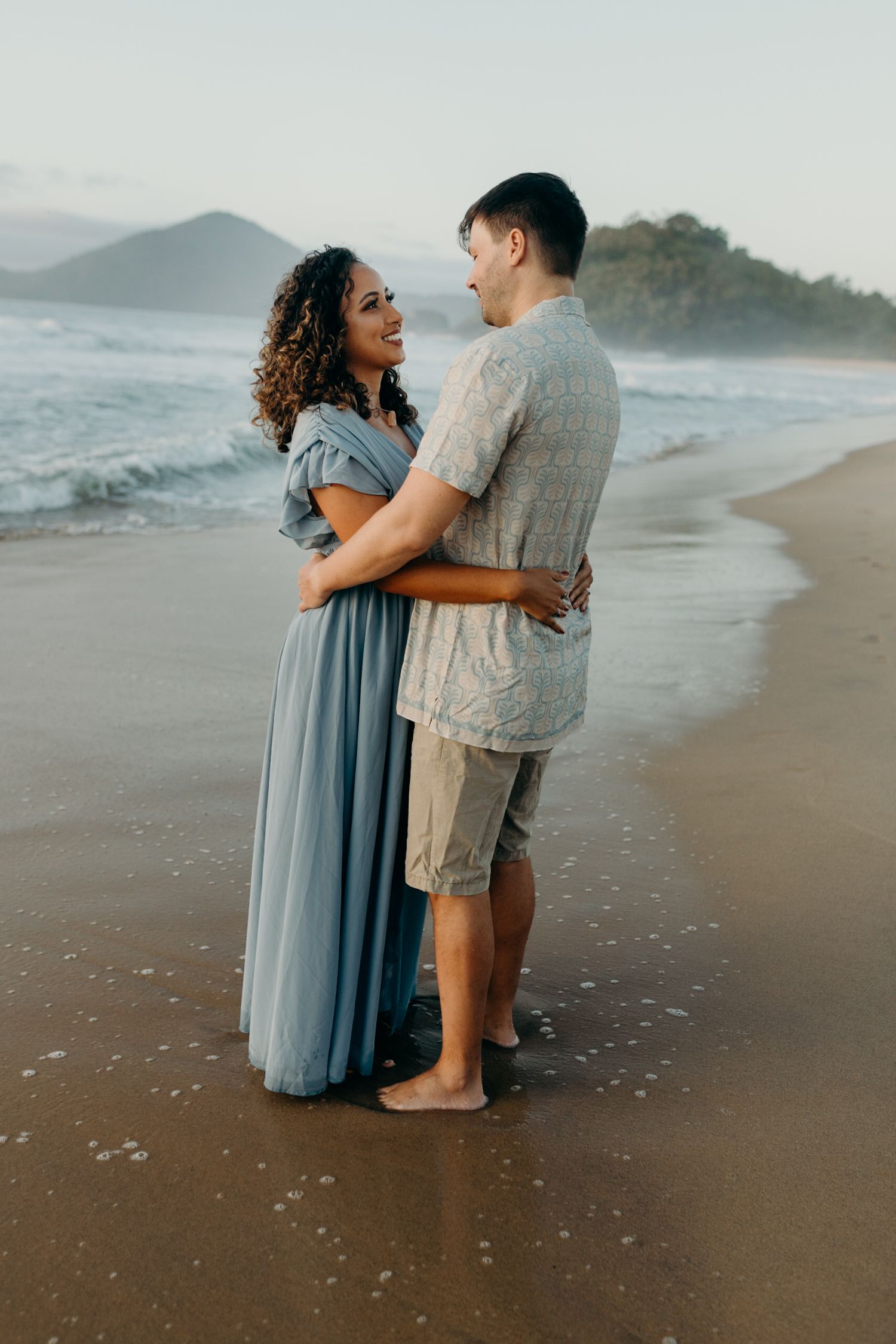 Couple wading in shallow ocean waves during beach photoshoot with mountains in distance.