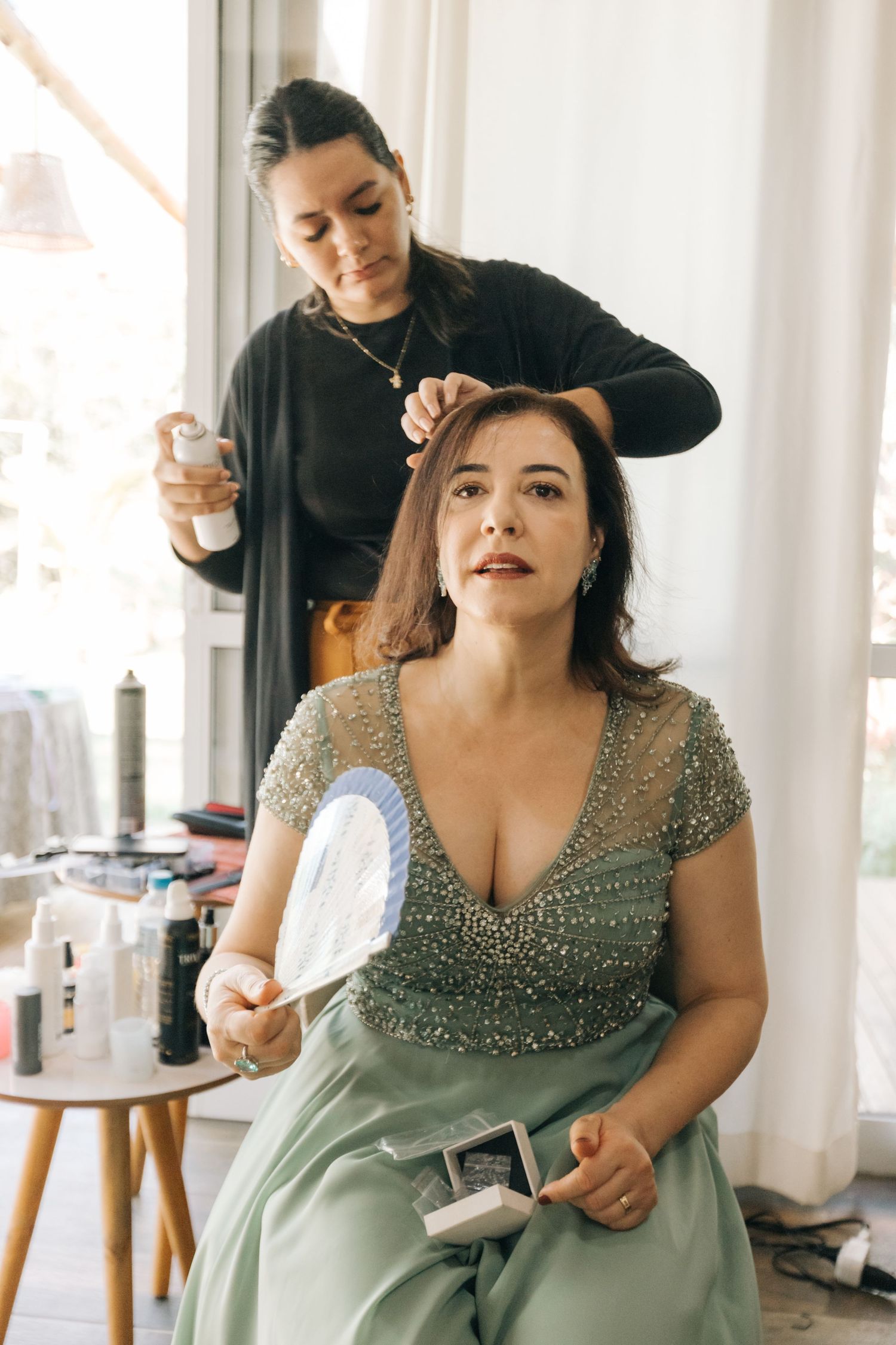 Woman in a sparkly sage green dress getting her makeup done in a bridal preparation room.