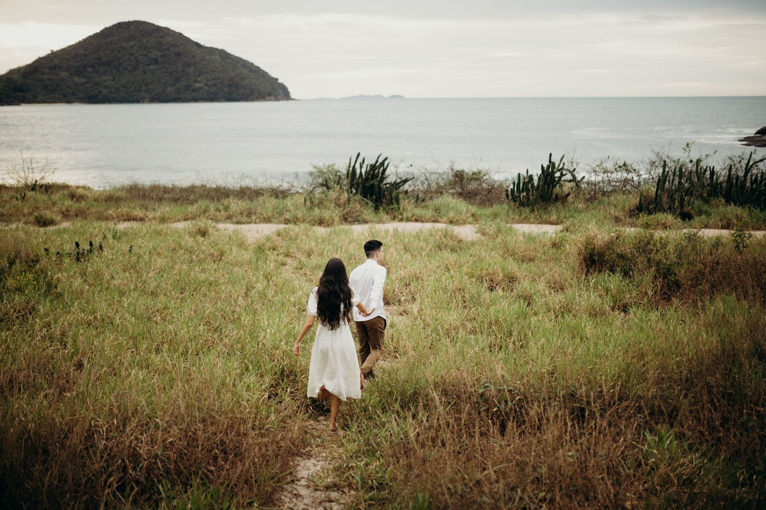 Couple walks hand in hand through coastal grasslands with ocean view.