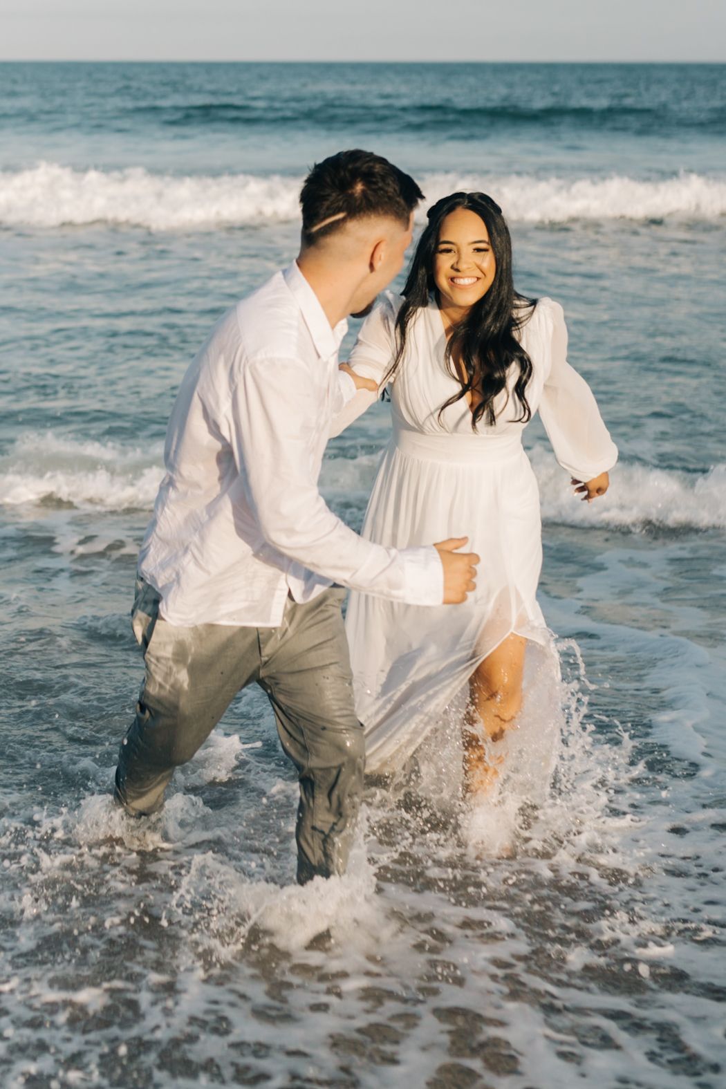 Playful couple runs through ocean waves on a beach while wearing white clothing and laughing together.
