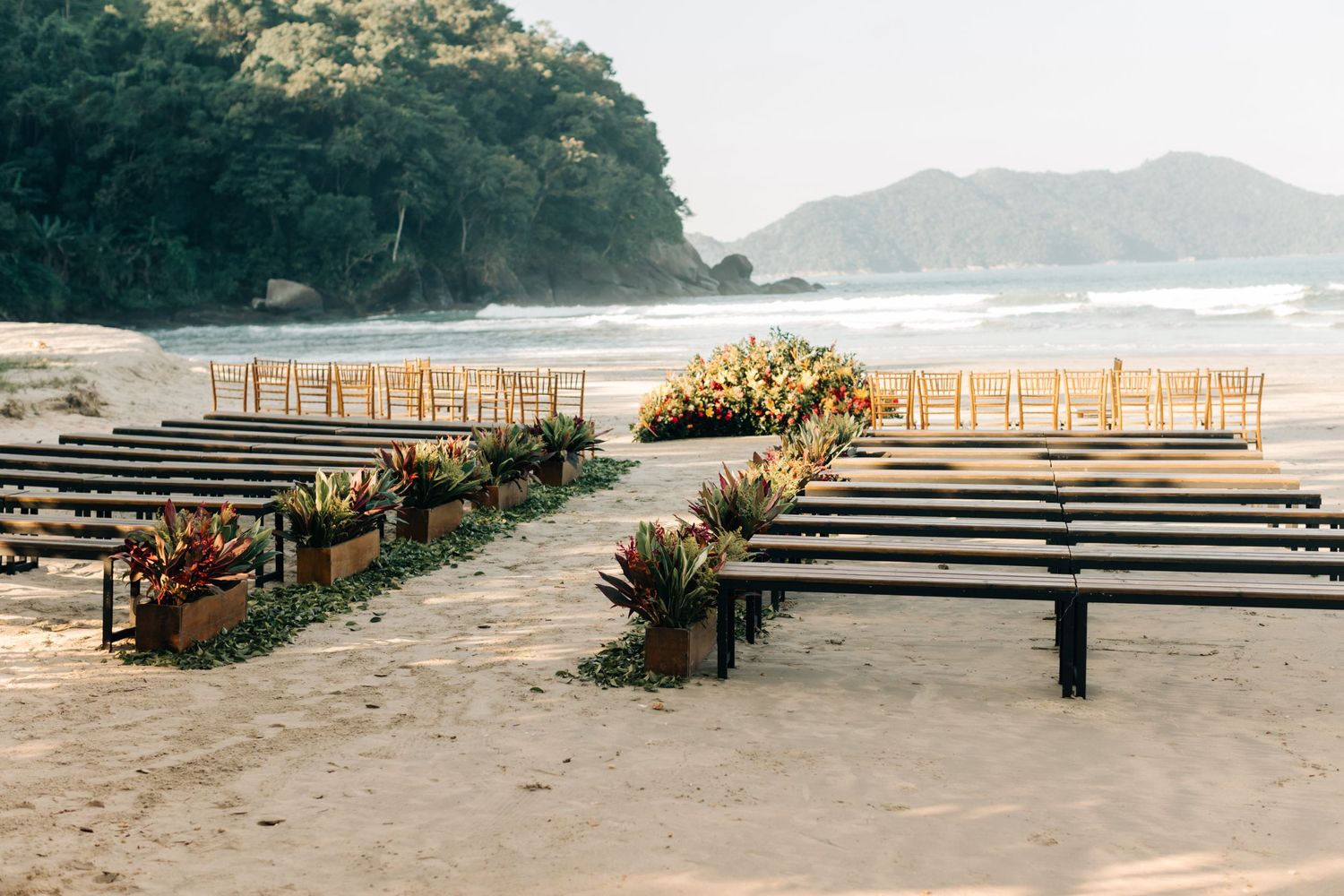 Wedding ceremony site on beach with rows of benches and tropical floral arrangements.
