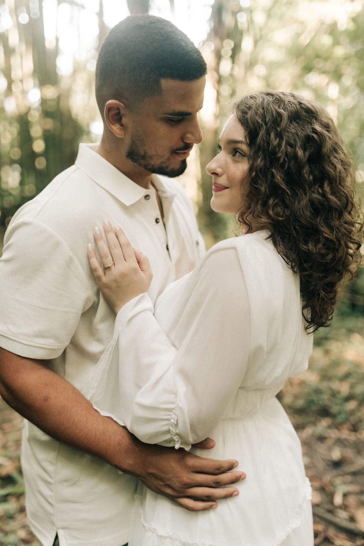 Loving couple shares tender moments during forest photoshoot wearing matching white outfits.