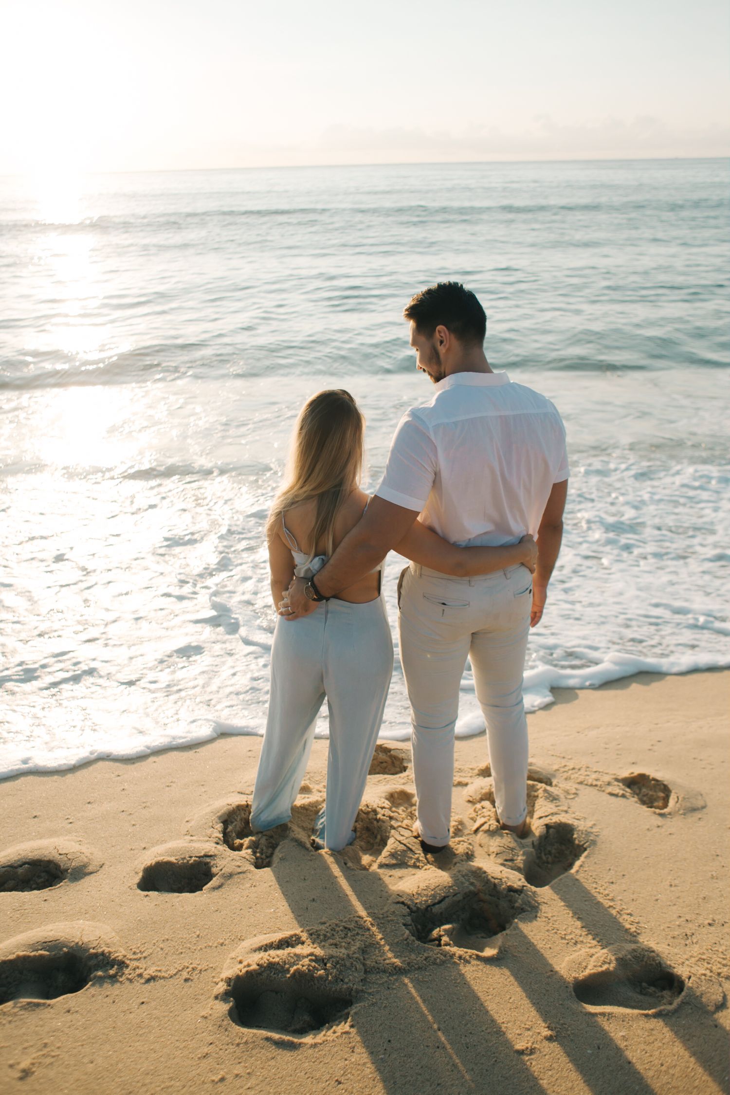 Two people in white outfits standing together watching ocean waves at sunset.