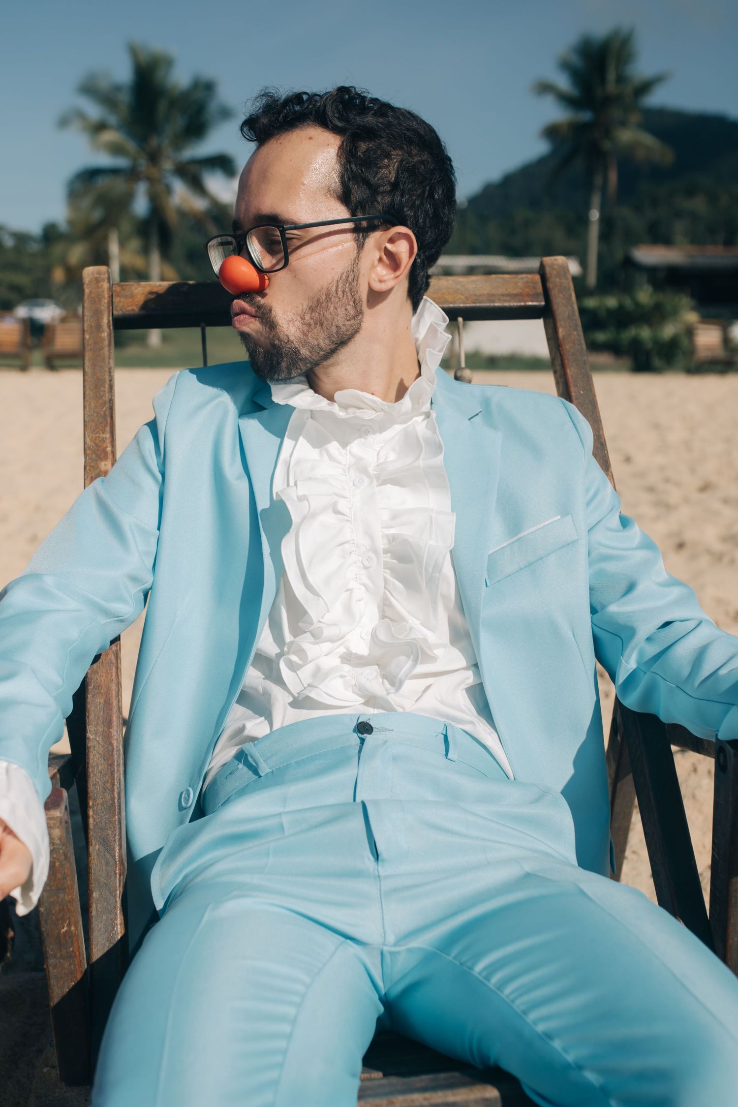 Person in light blue suit and white shirt lounging in beach chair with red nose.