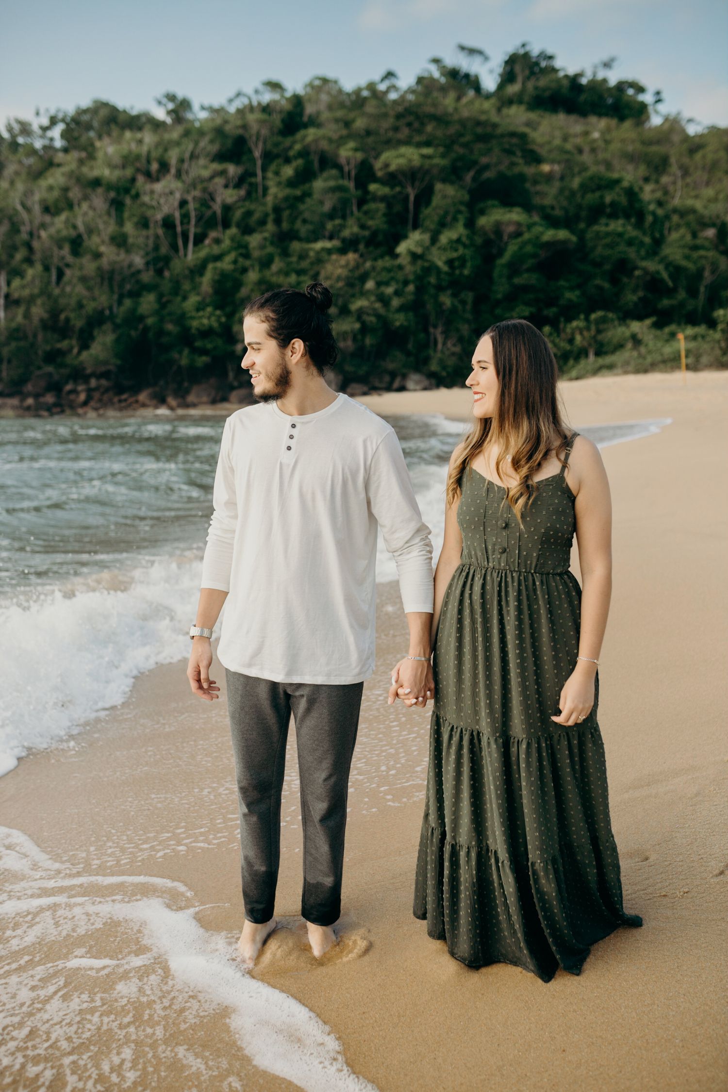 A couple walks barefoot along a scenic beach with forest-covered cliffs in the background.