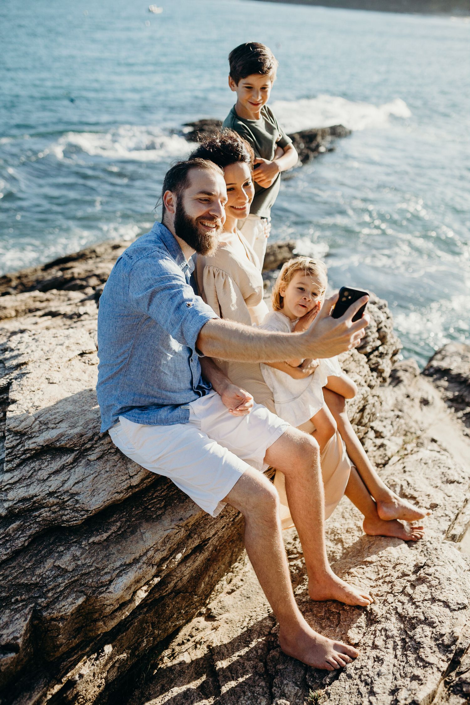 Family shares intimate moment while sitting on rocky coastal outcrop with waves crashing below.