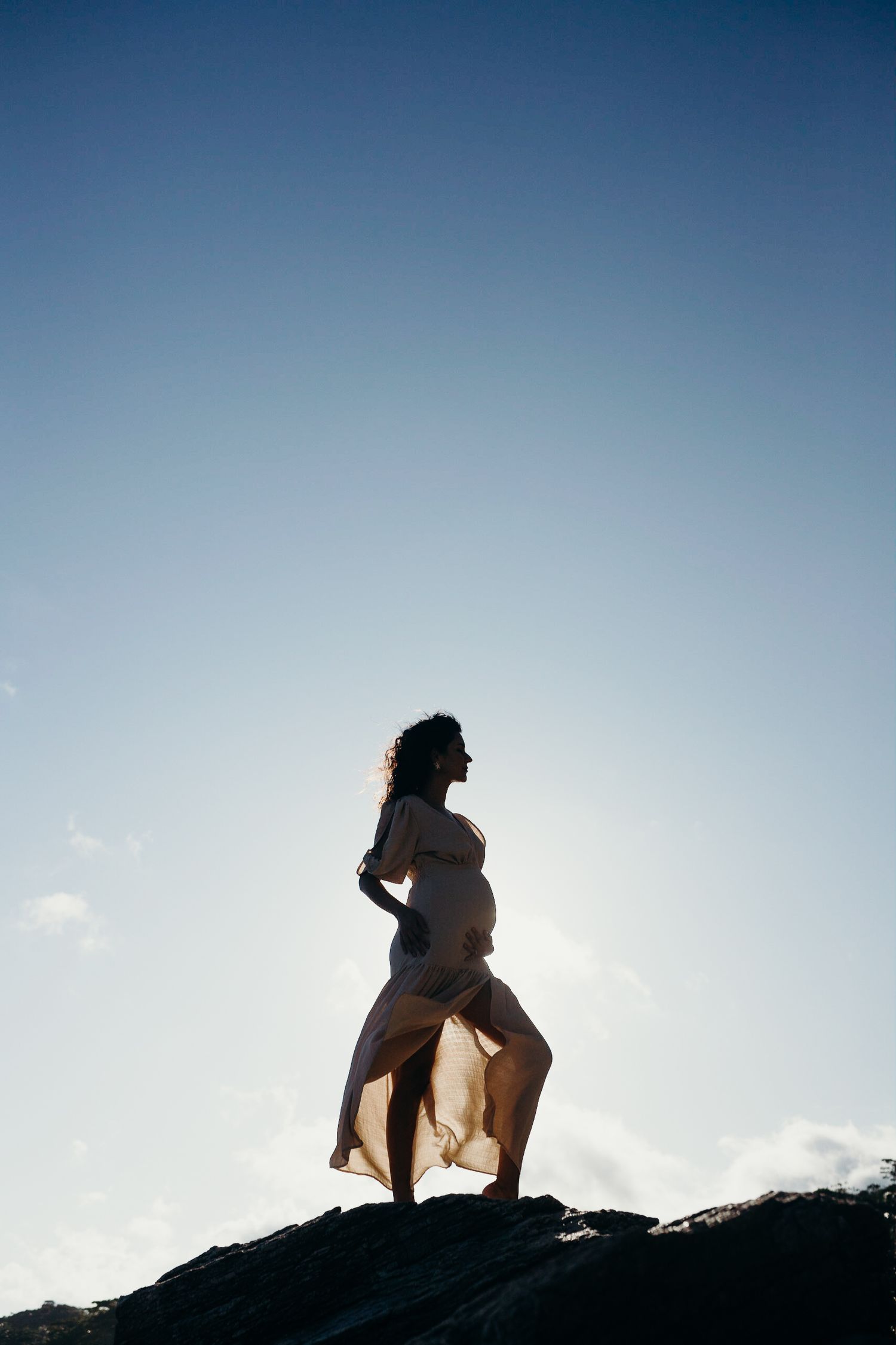 Maternity silhouette standing on coastal rocks with dress flowing in wind.