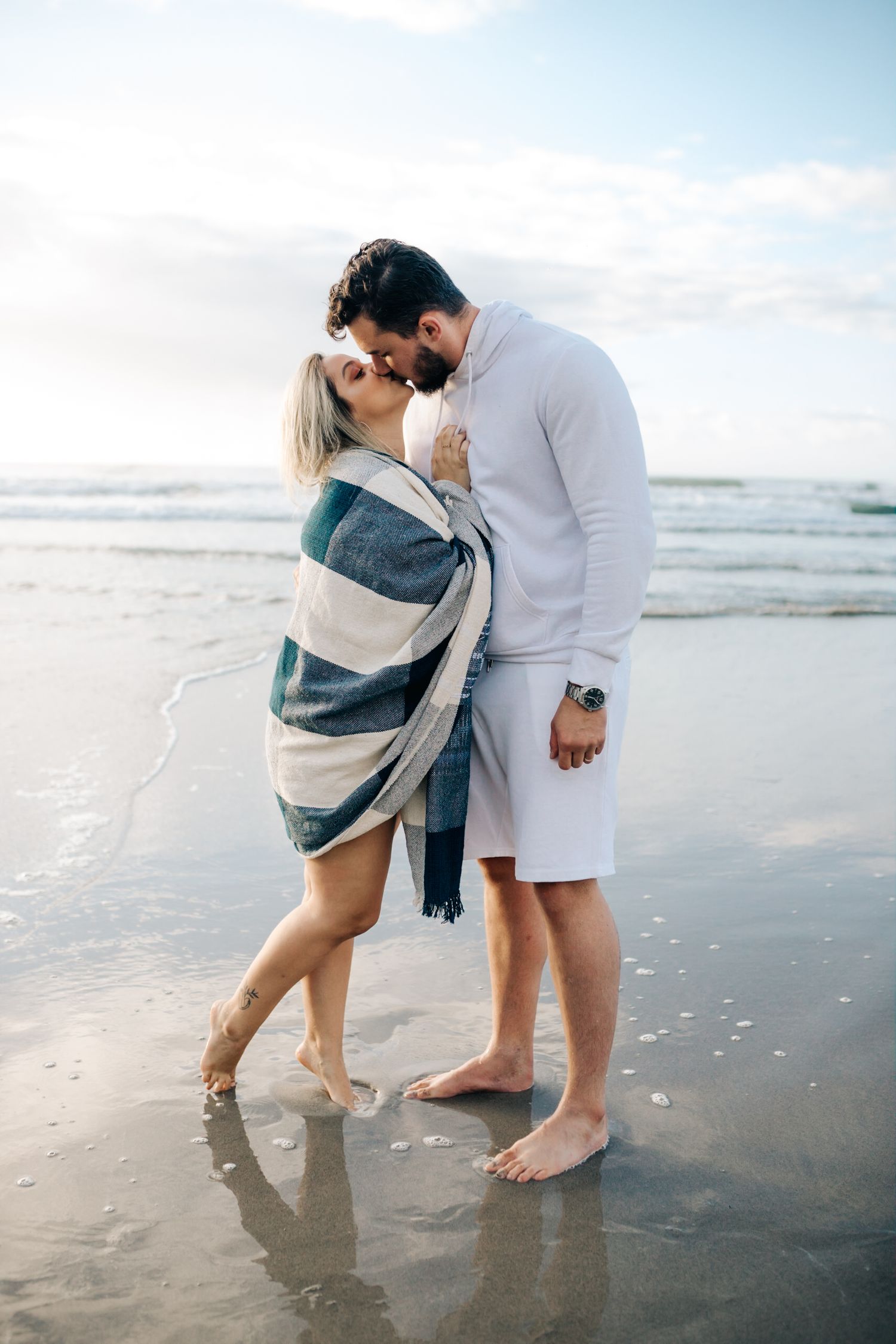 Romantic embrace between couple standing barefoot on wet sand at the ocean's edge.