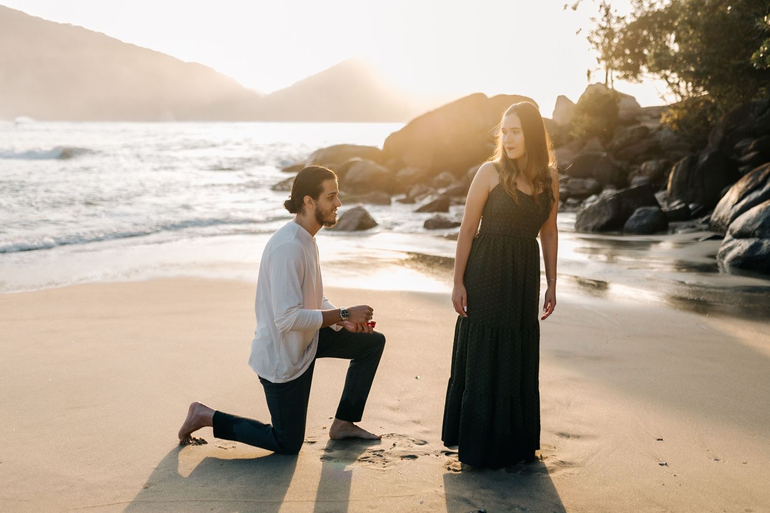 A romantic beach proposal during golden hour with waves and mountains in the distance.