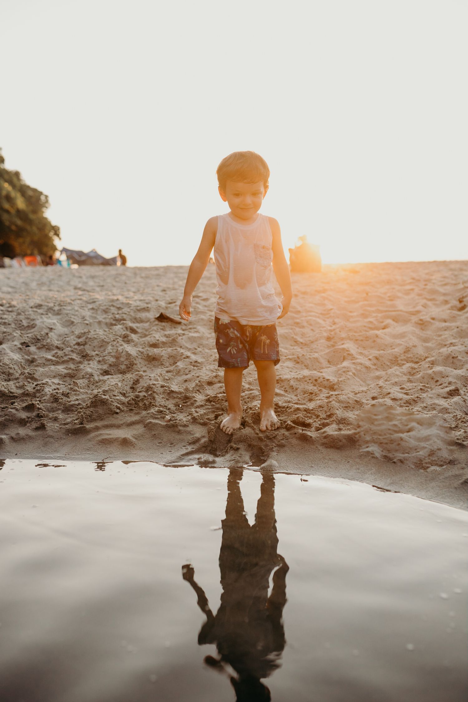 Small child standing alone at water's edge during sunset, creating reflection in shallow water.