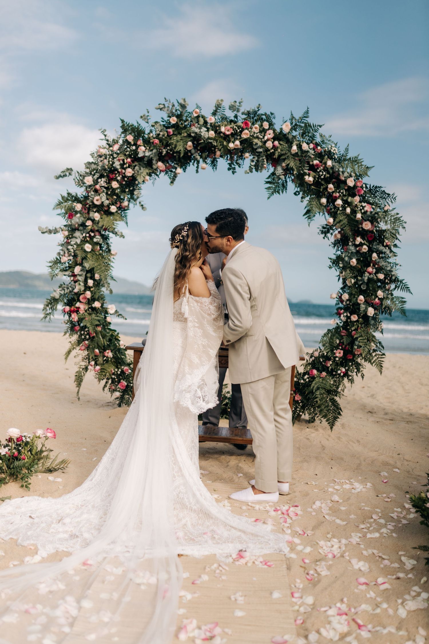 A wedding couple shares their first kiss under a beautiful floral arch on the beach.
