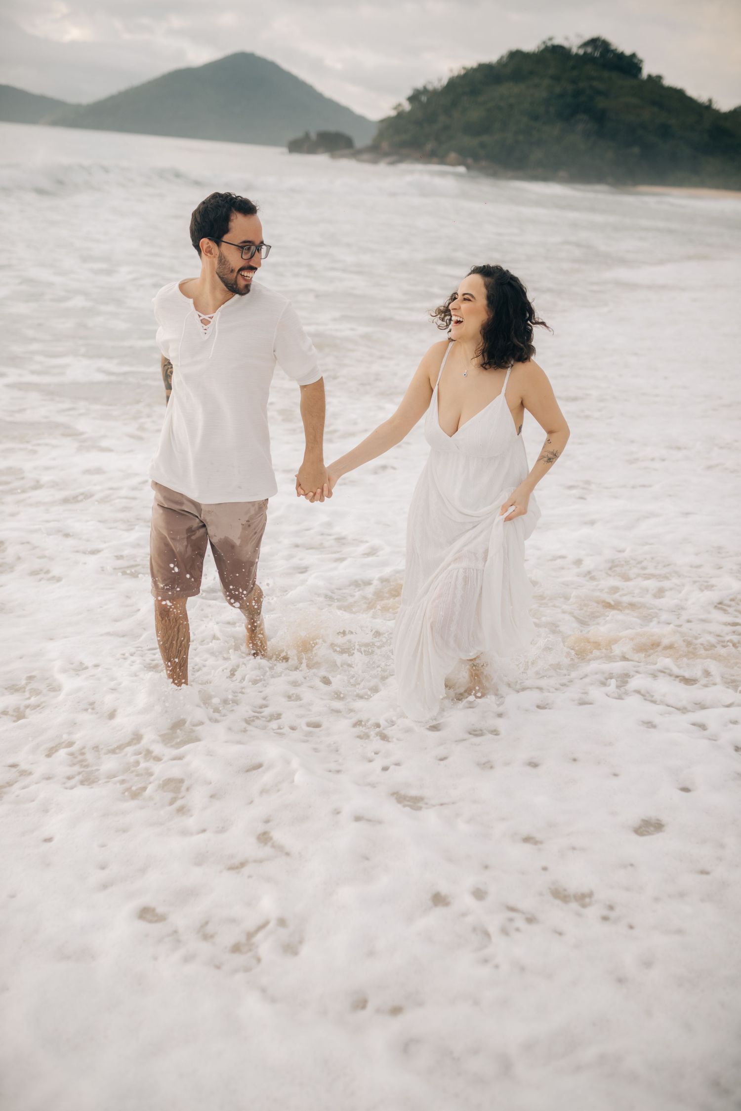 A couple wades through ocean foam while holding hands on a beach with mountains in the distance.