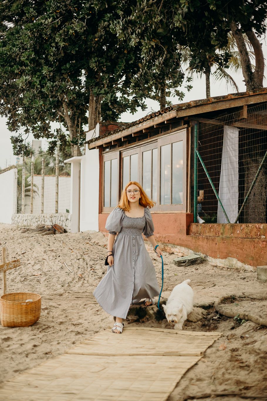 Woman in a gray maxi dress strolls on a wooden walkway with a white dog.
