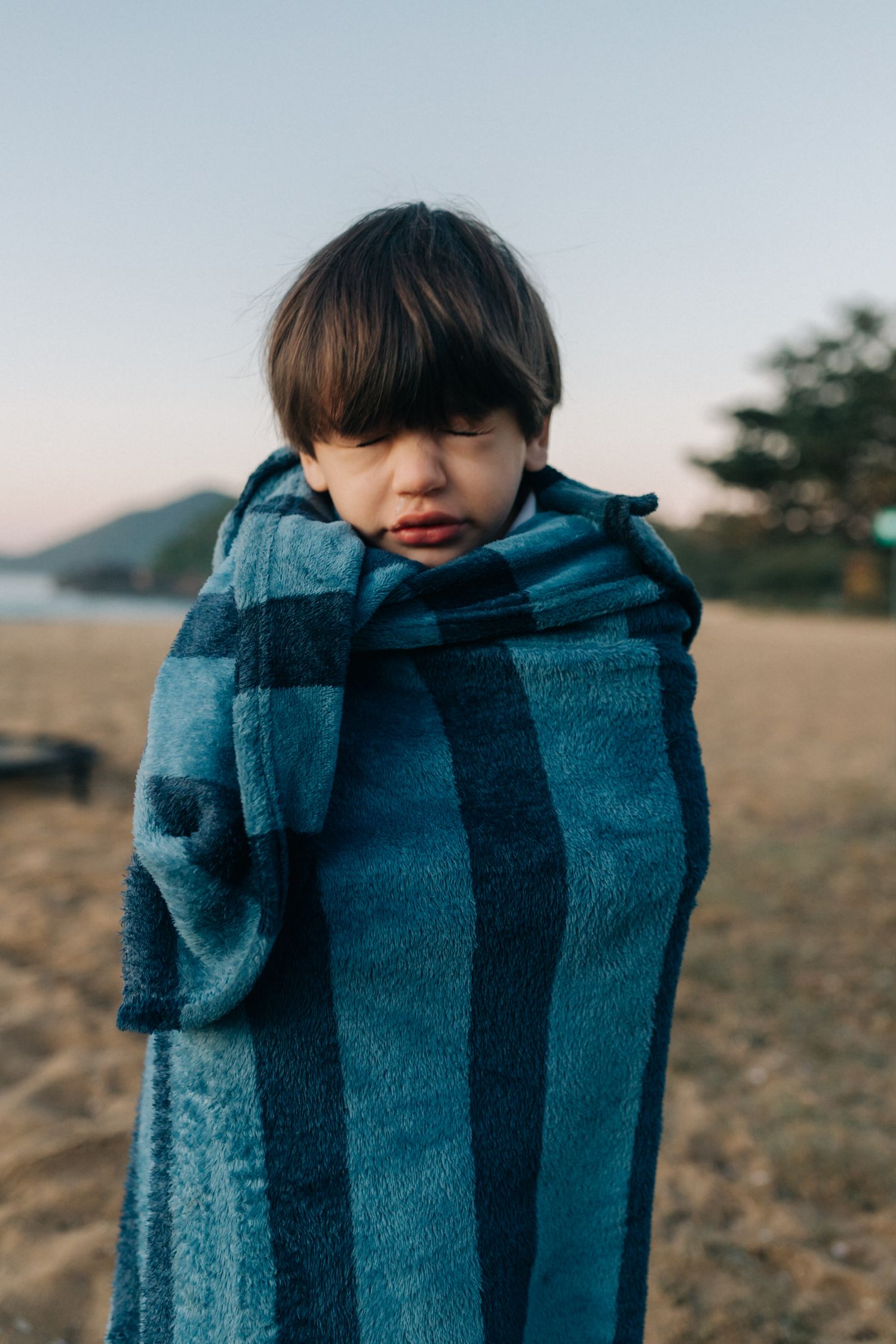 Young person bundled in blue striped blanket against coastal backdrop.