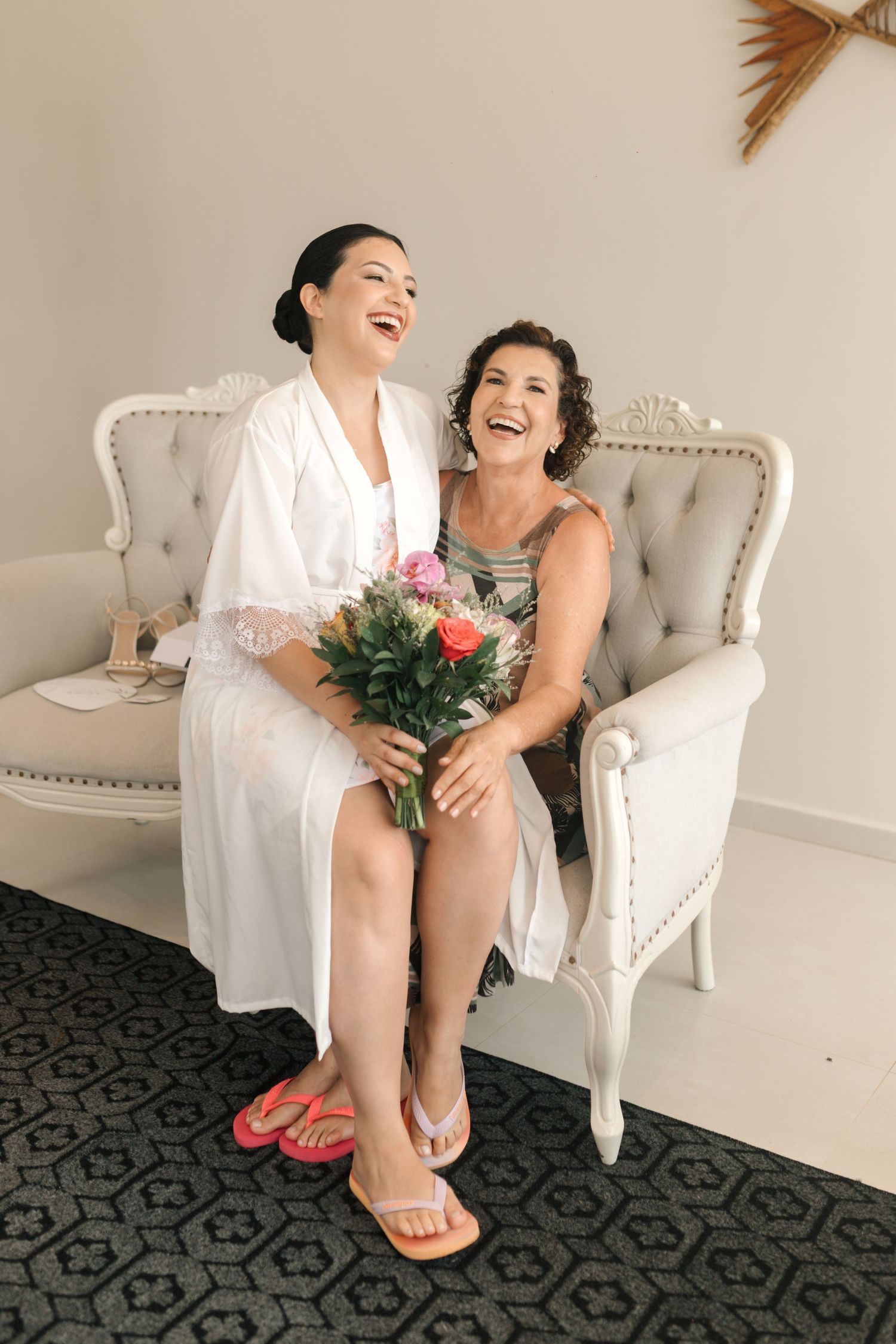 Two people share joyful laughter while sitting on an elegant white tufted loveseat holding a pink floral bouquet.