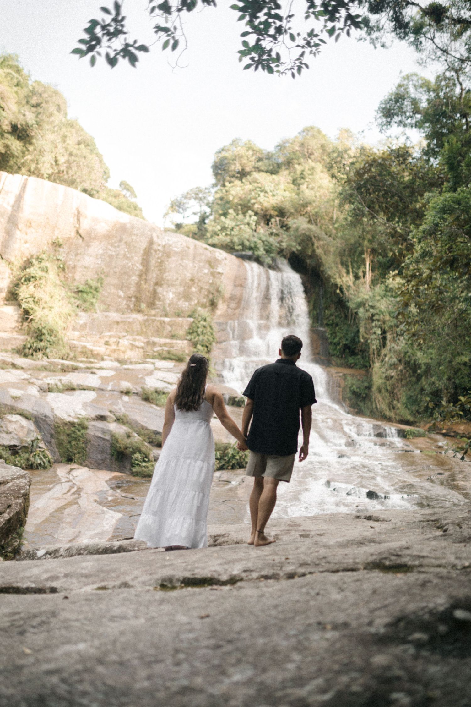 Couple walking hand in hand near scenic waterfall in tropical setting.