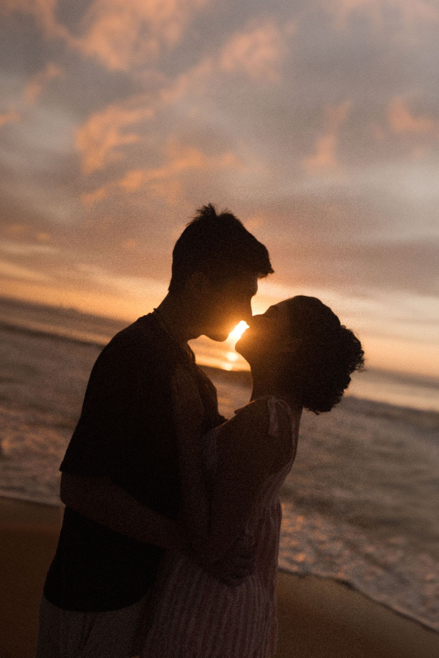 A silhueta de um casal se abraça em uma praia ao pôr do sol com nuvens douradas e ondas do mar ao fundo.